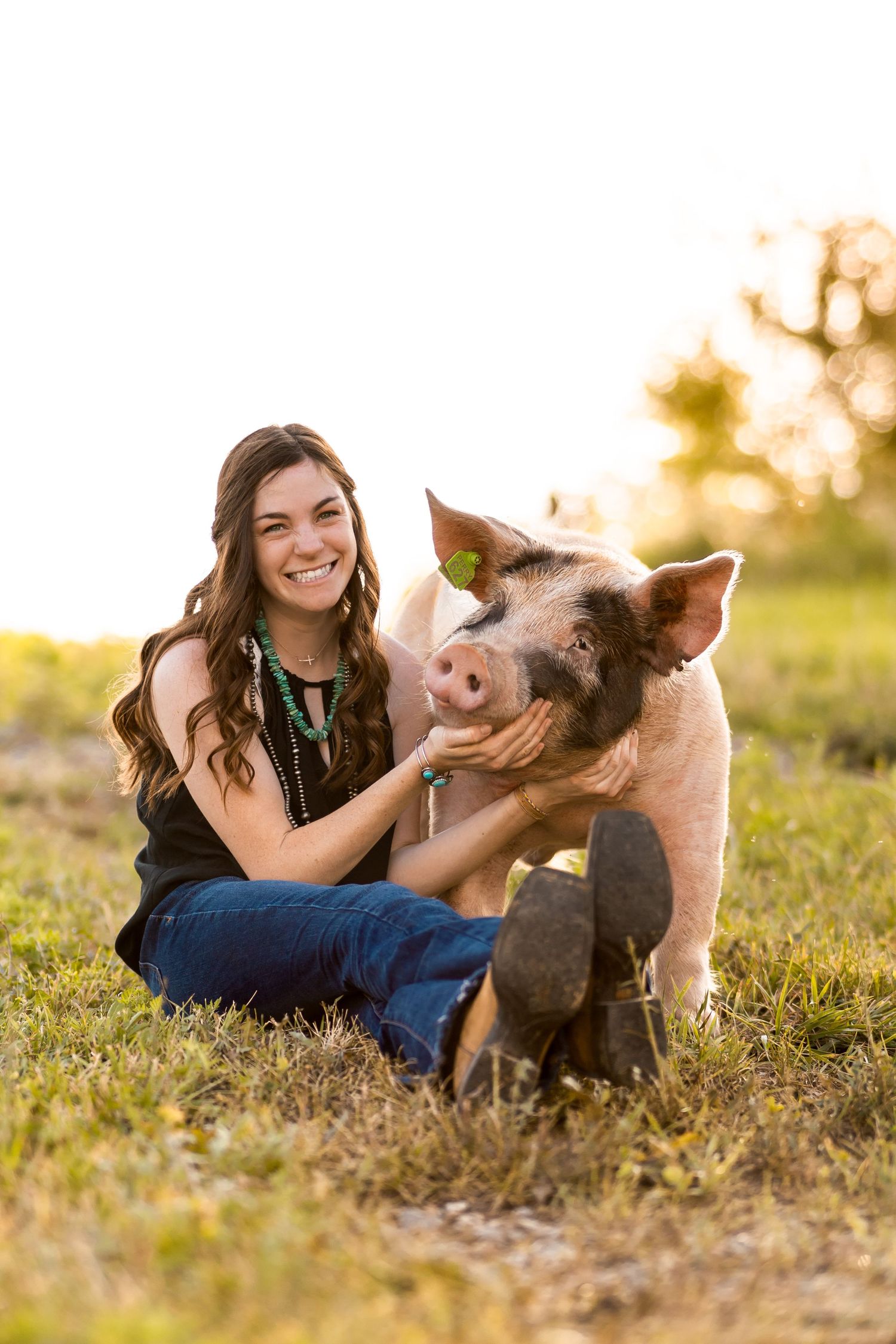 Young woman smiling and petting a pig while sitting in a sunny green field, wearing jeans and a turquoise necklace.