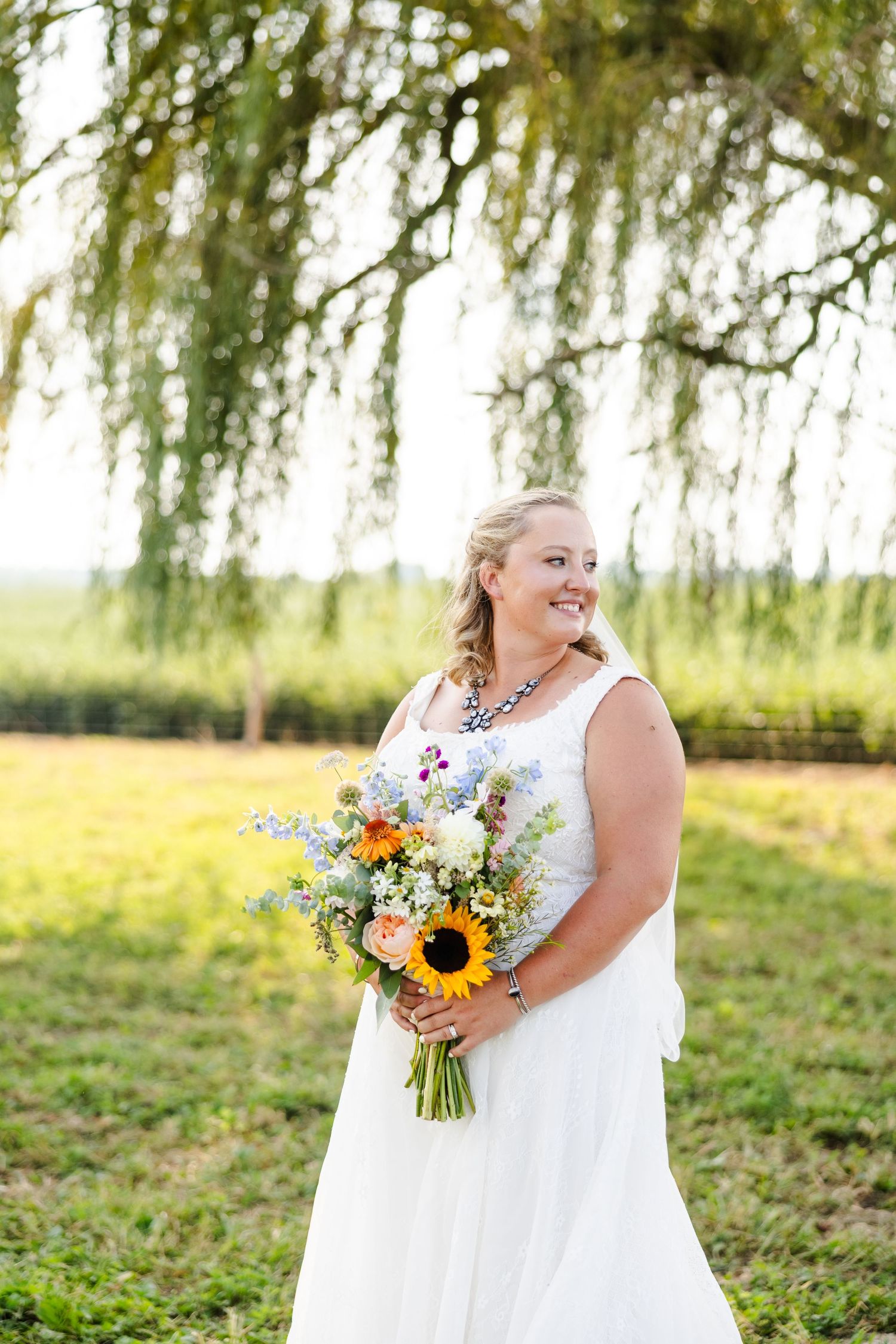 Bride in a white gown smiles while holding a colorful sunflower and wildflower bouquet under a weeping willow.