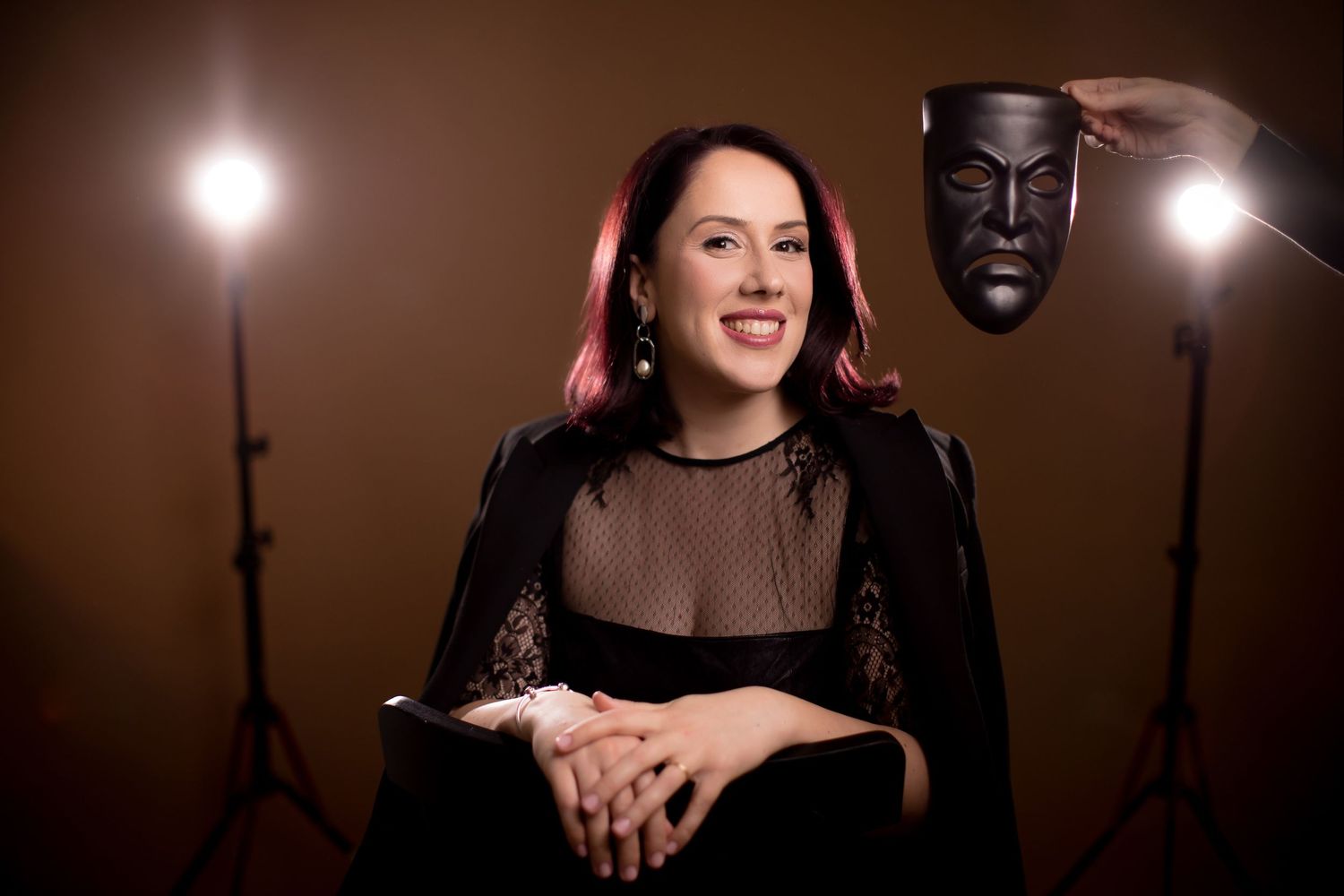 Woman in black dress smiling in studio with stage lights and a dramatic theater mask held beside her.