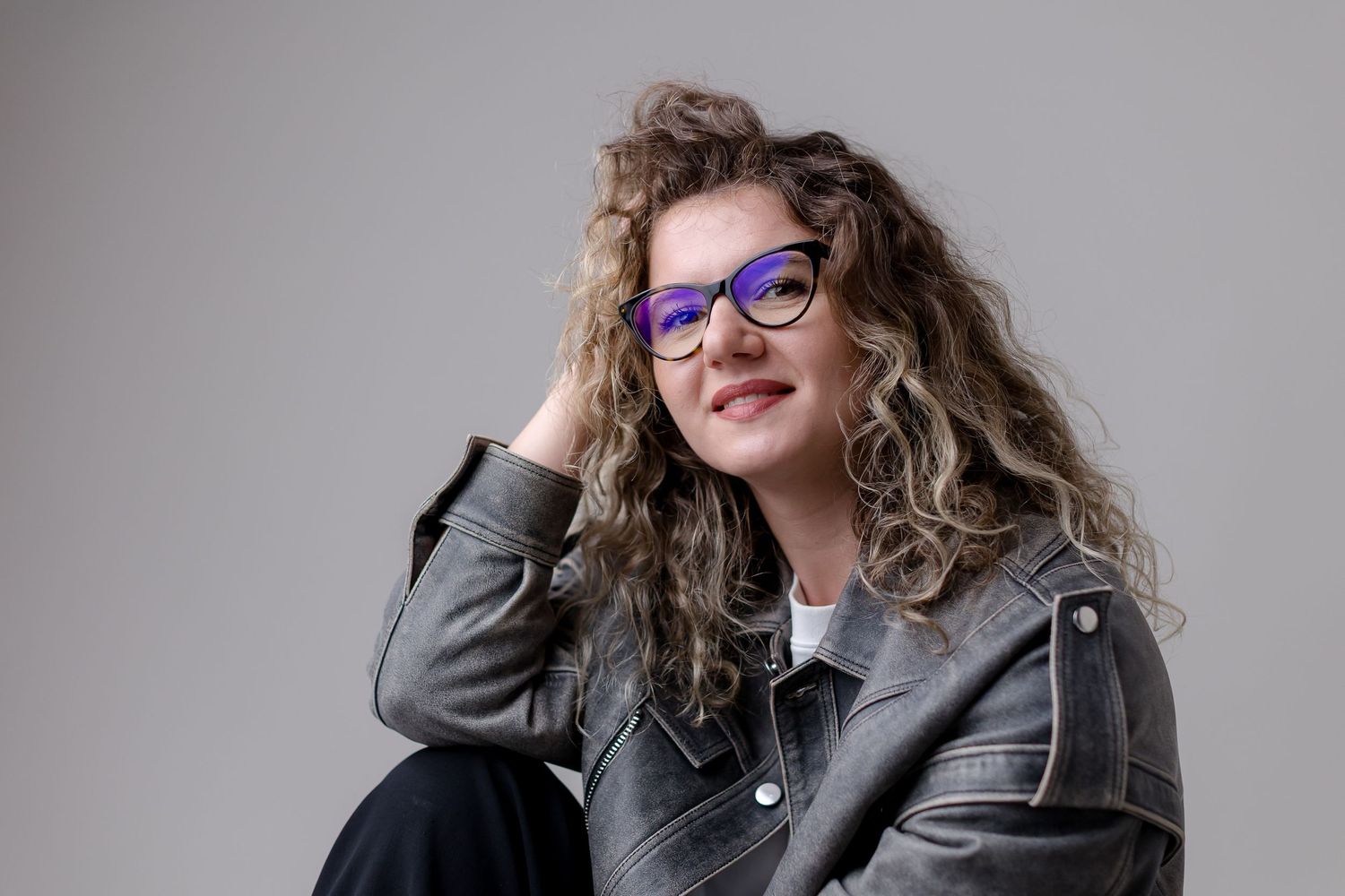 Young woman with curly hair and purple glasses wearing a denim jacket, smiling in a studio portrait.