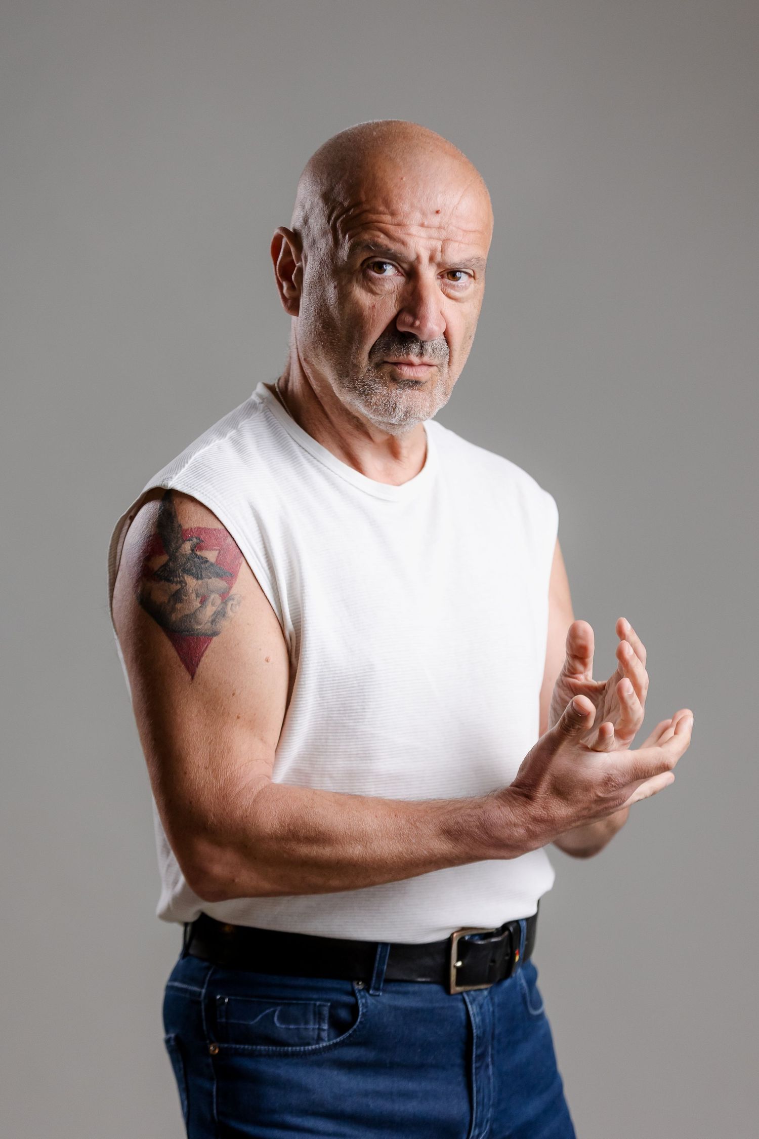 Bald middle-aged man with arm tattoo in white sleeveless shirt and jeans gesturing with hands against gray background.