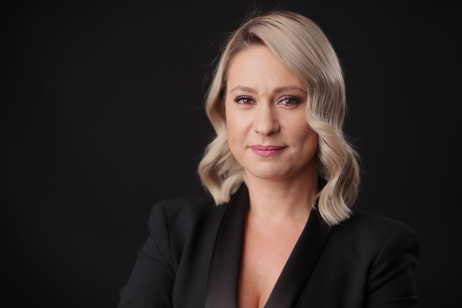Professional headshot of a blonde woman in a black blazer against a dark studio background.
