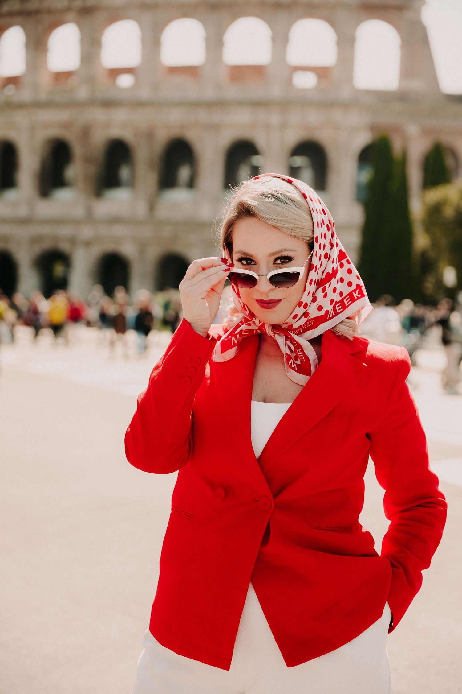Stylish blonde woman in a red blazer and headscarf adjusting sunglasses in front of the Roman Colosseum.