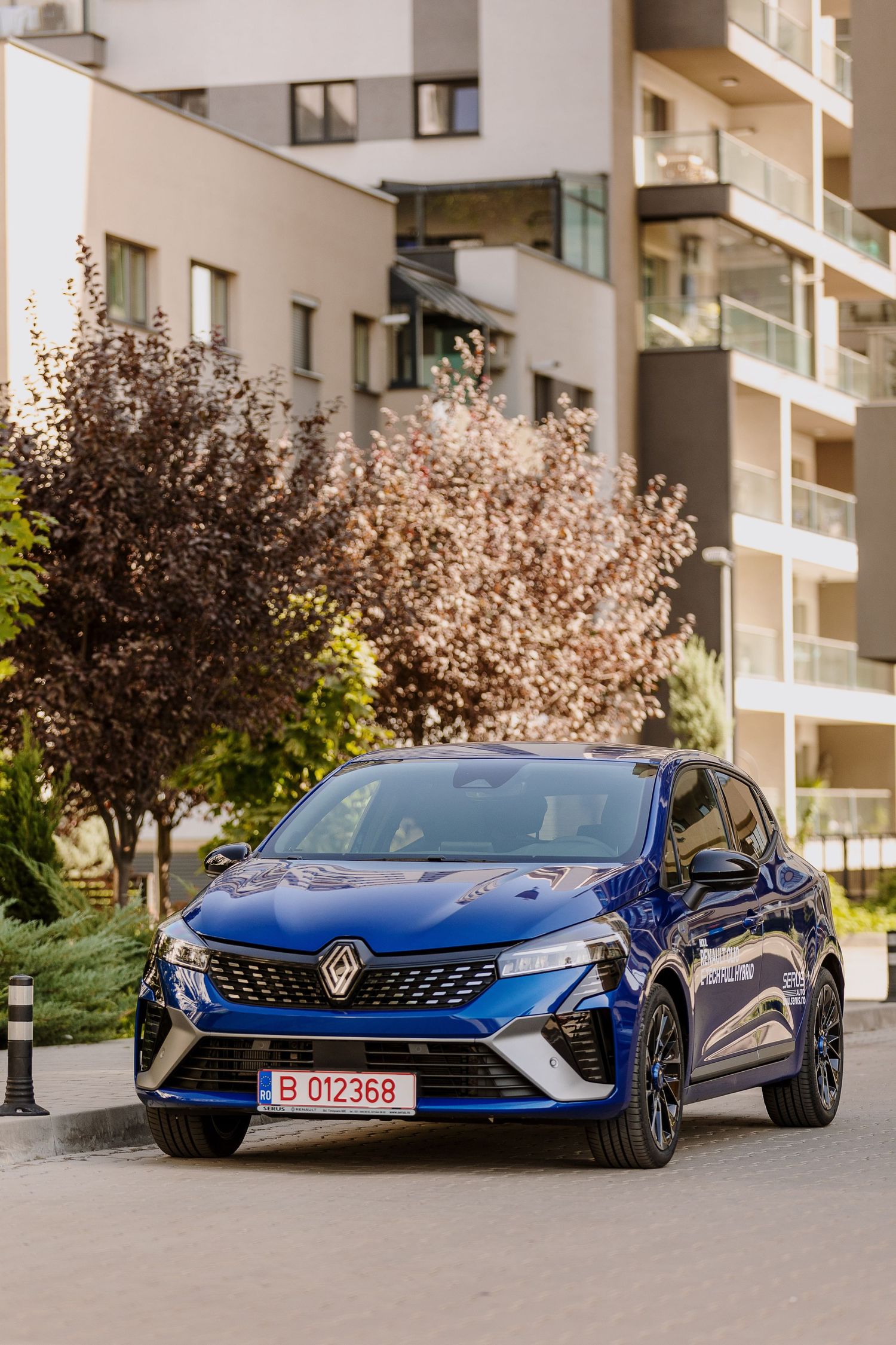 Blue Renault hatchback parked in modern residential area with decorative trees in background.