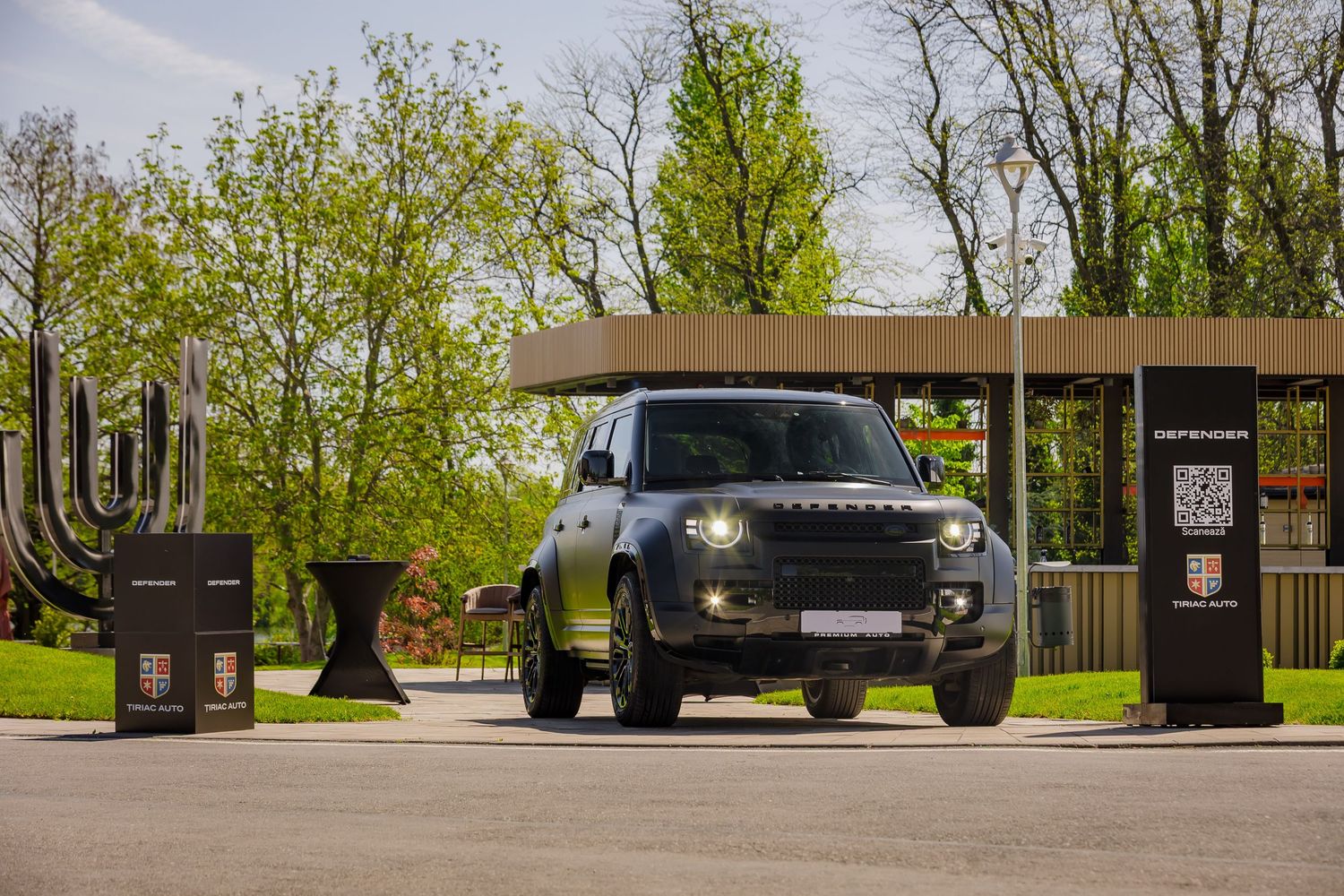 Dark Land Rover Defender SUV parked at an outdoor event entrance with green trees and wooden canopy in background.