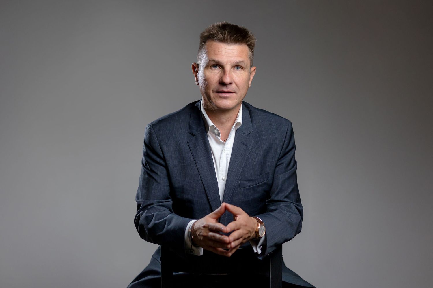 Professional businessman in blazer sitting confidently with hands clasped, studio portrait on gray background.