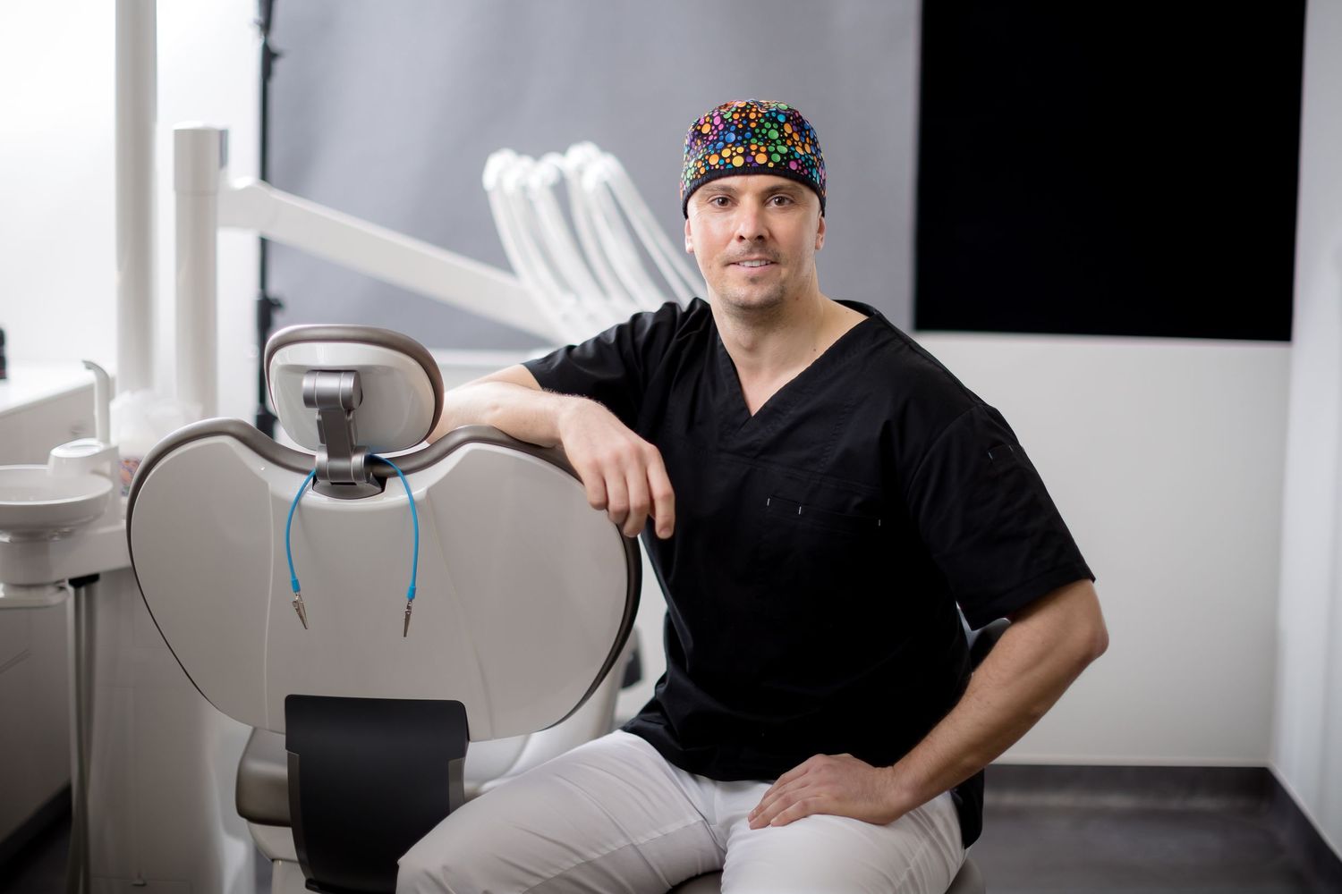 Male dentist in black scrubs and colorful cap poses confidently in modern dental office with equipment.