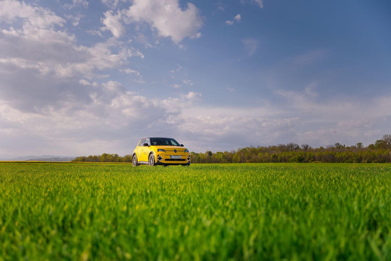 Yellow car parked in a lush green field under a blue sky with fluffy white clouds.