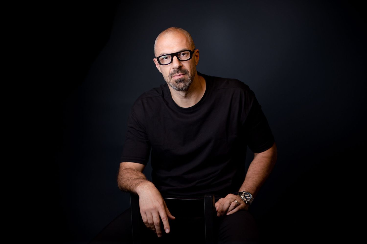 Bald man with glasses in black t-shirt poses confidently against dark background in professional portrait photo.