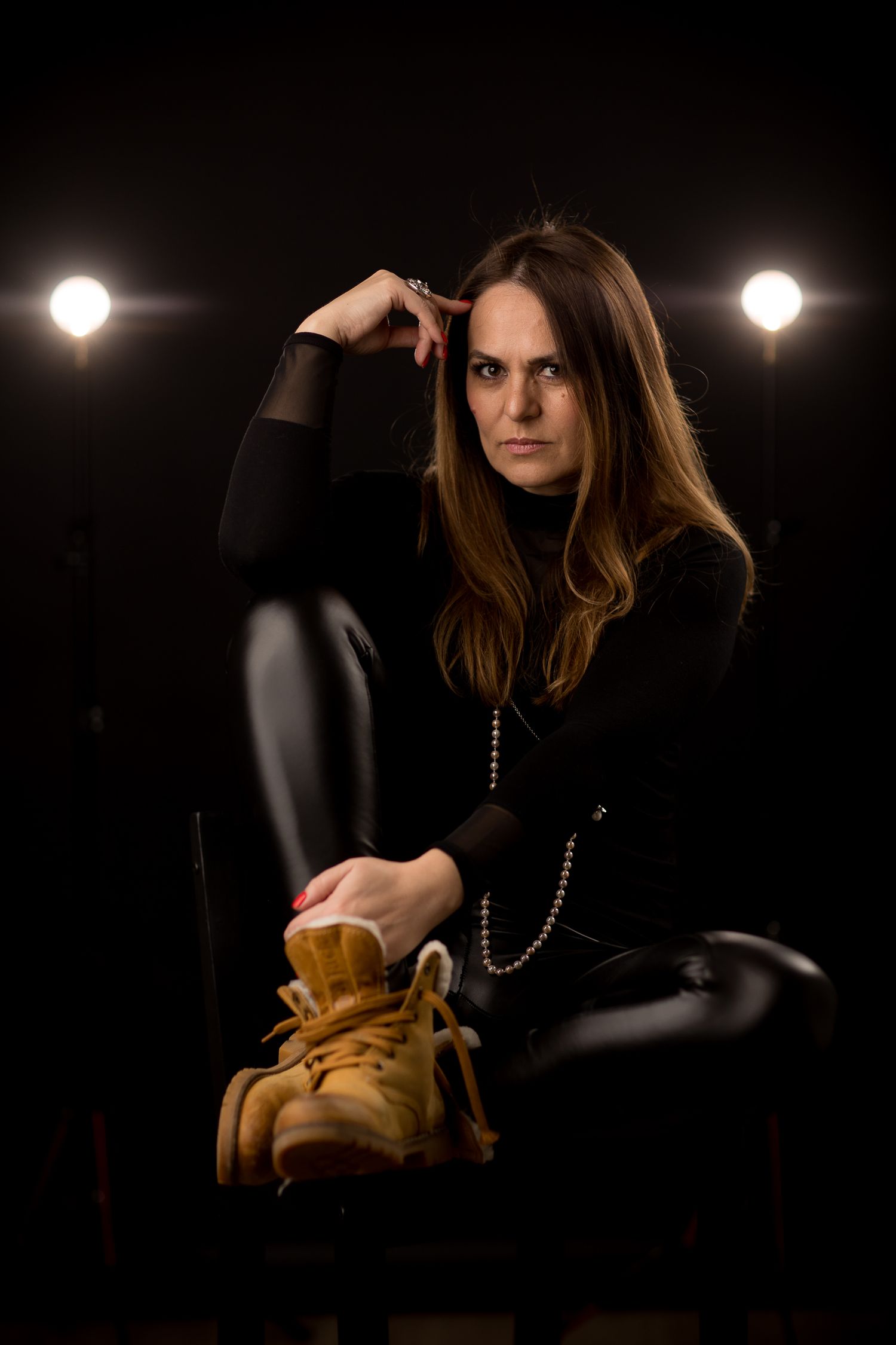 Woman in black outfit sitting on chair holding tan boots, posed against dark background with studio lights.