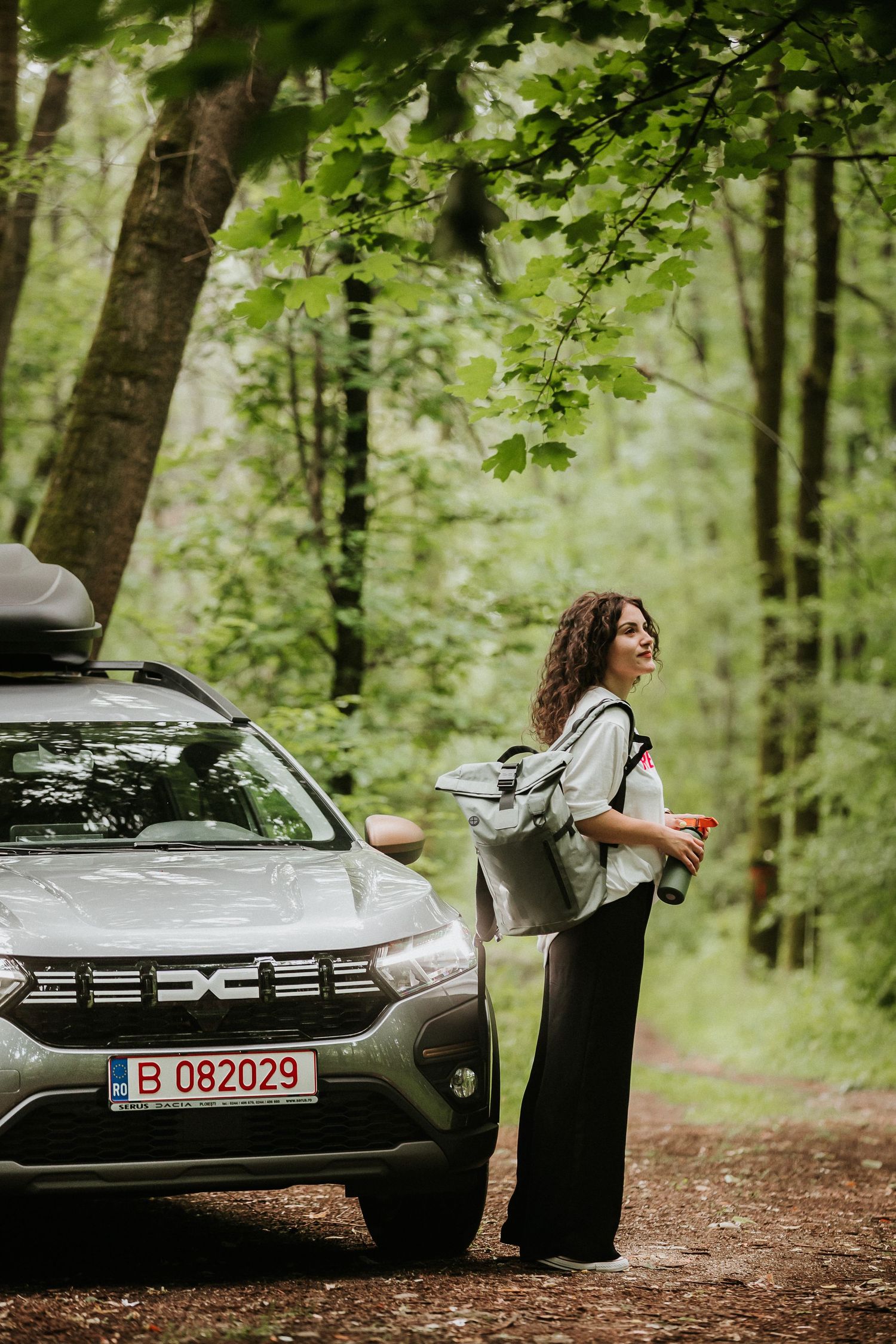Woman with backpack standing beside a Dacia SUV on a forest trail surrounded by lush green trees.
