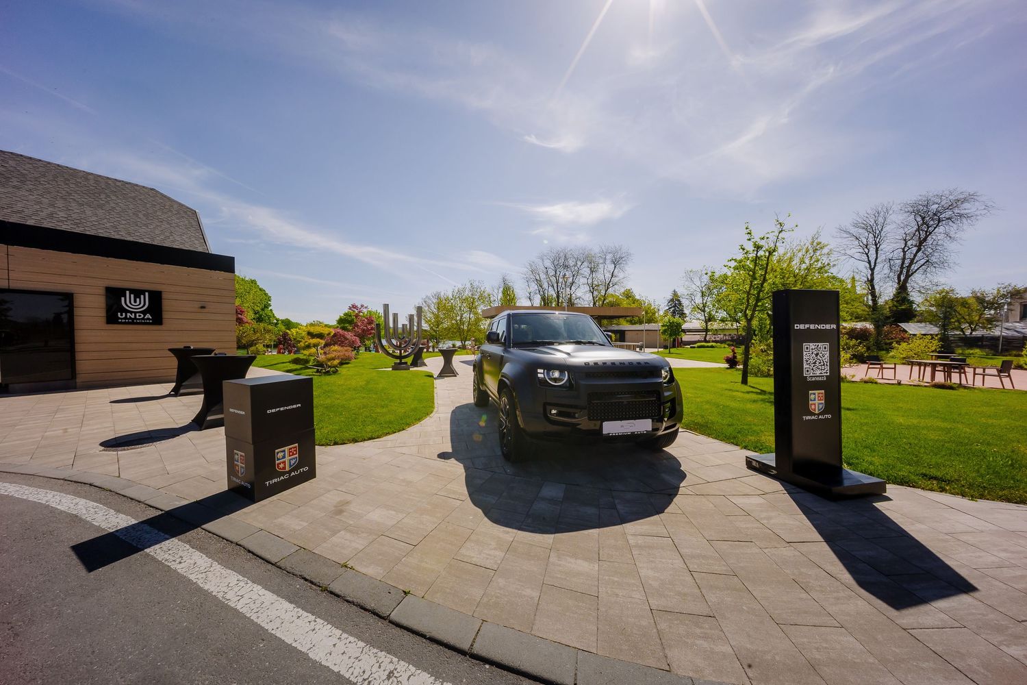 Dark SUV entering an elegant estate driveway with manicured gardens and a modern building on a sunny day.