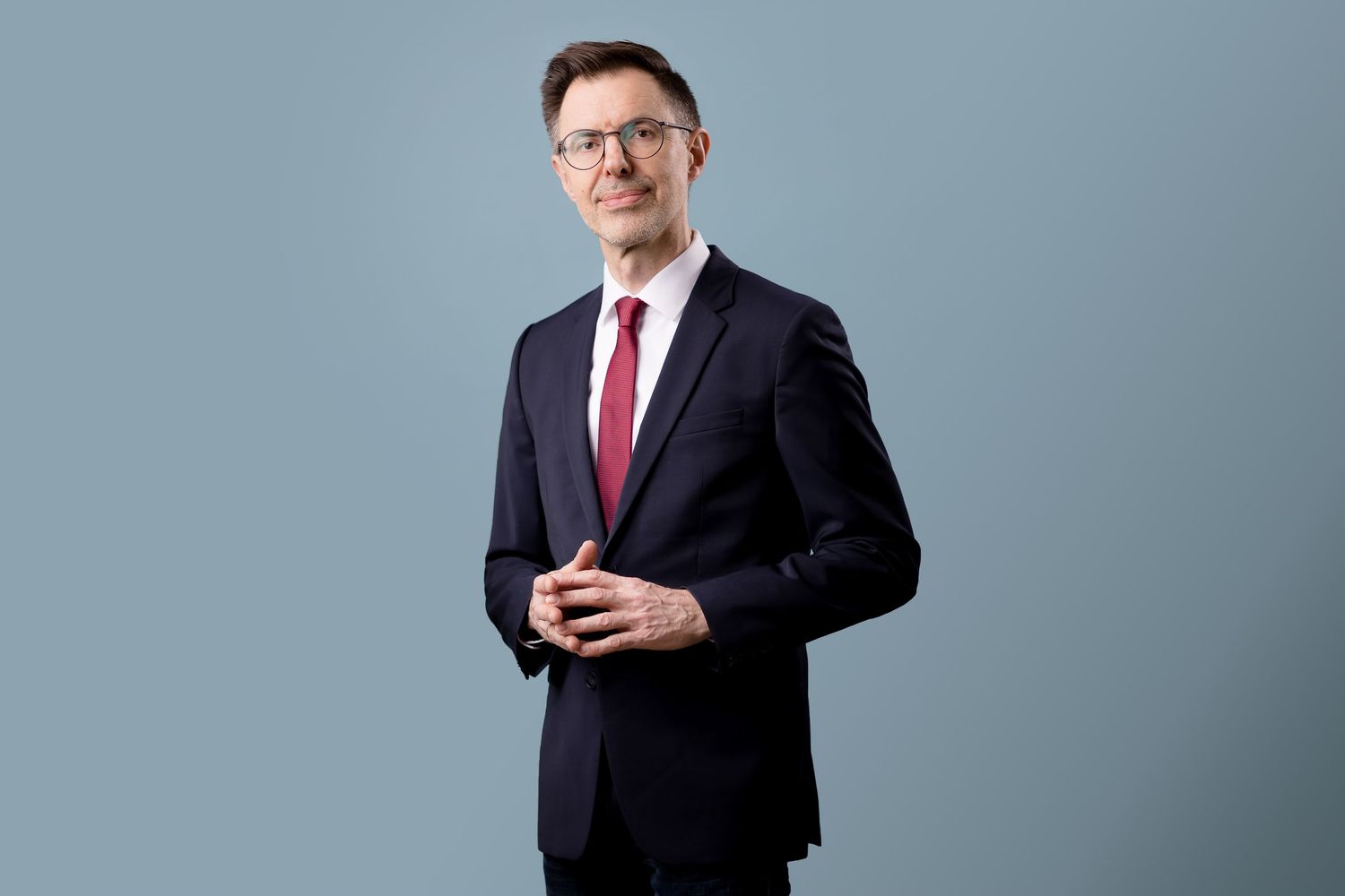 Professional businessman in navy suit and red tie smiling confidently against a gray background.