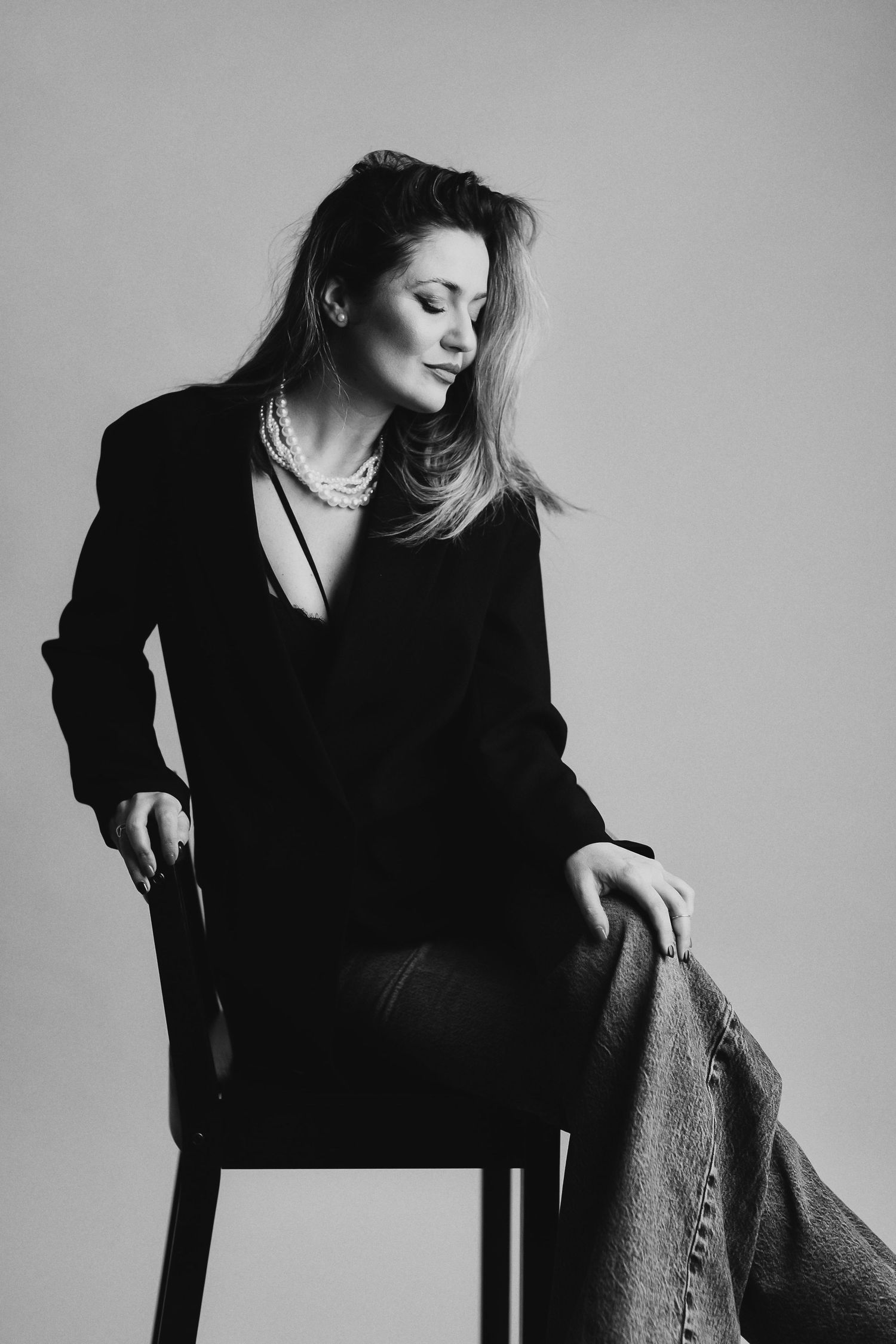 Black and white portrait of a woman in a black blazer and pearl necklace seated on a chair in a studio.
