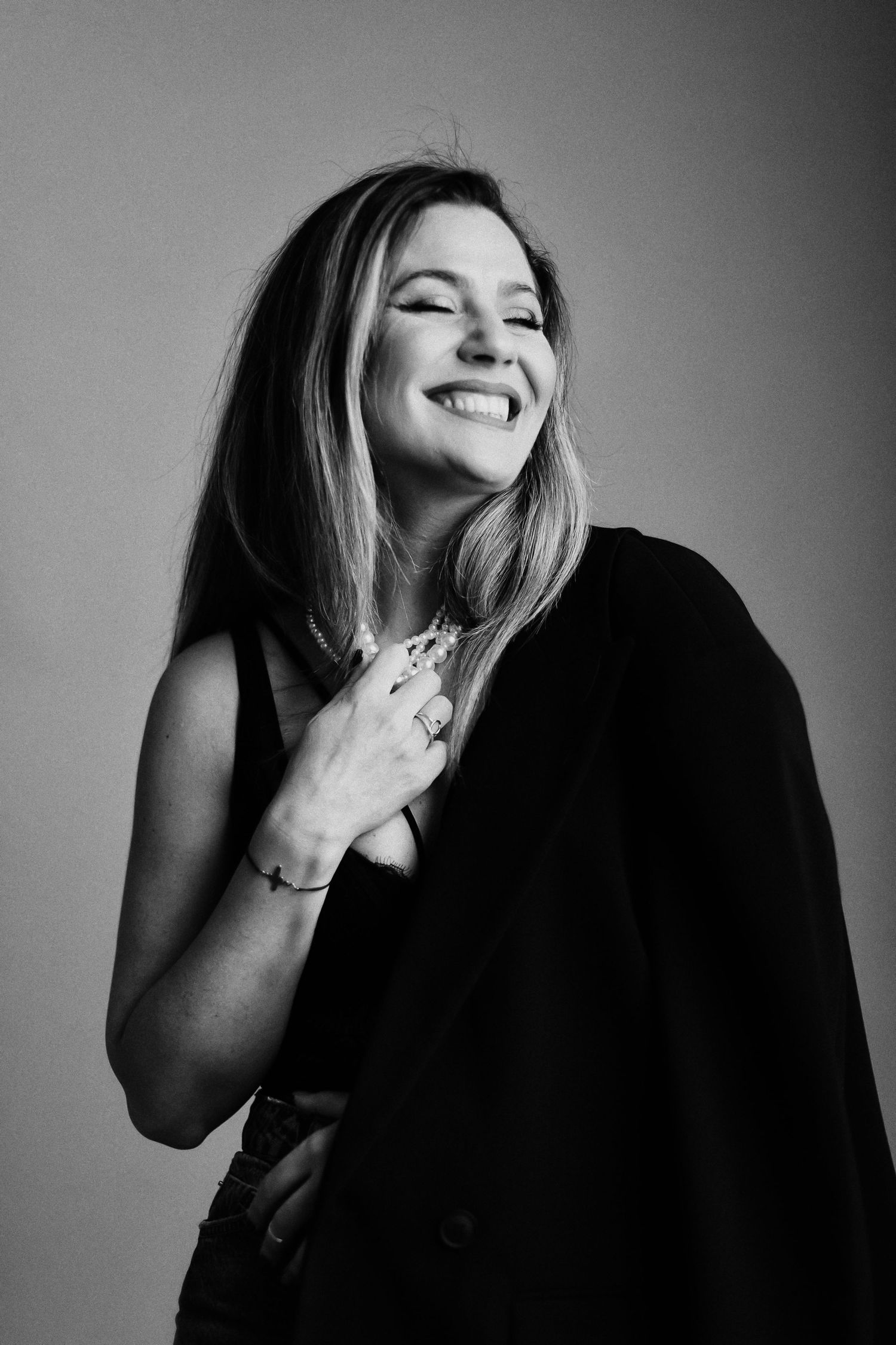 Black and white portrait of a smiling woman in a black blazer, laughing joyfully with hand on chest.