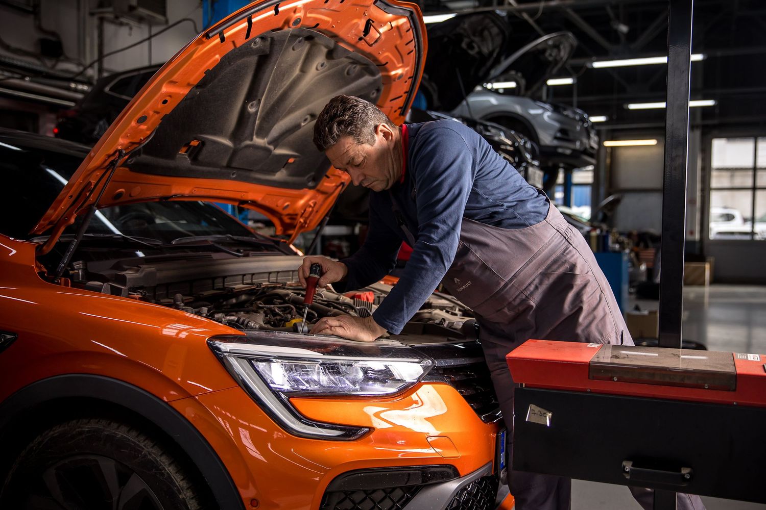 Mechanic in overalls inspecting engine of orange sports car with hood open in professional auto repair shop.