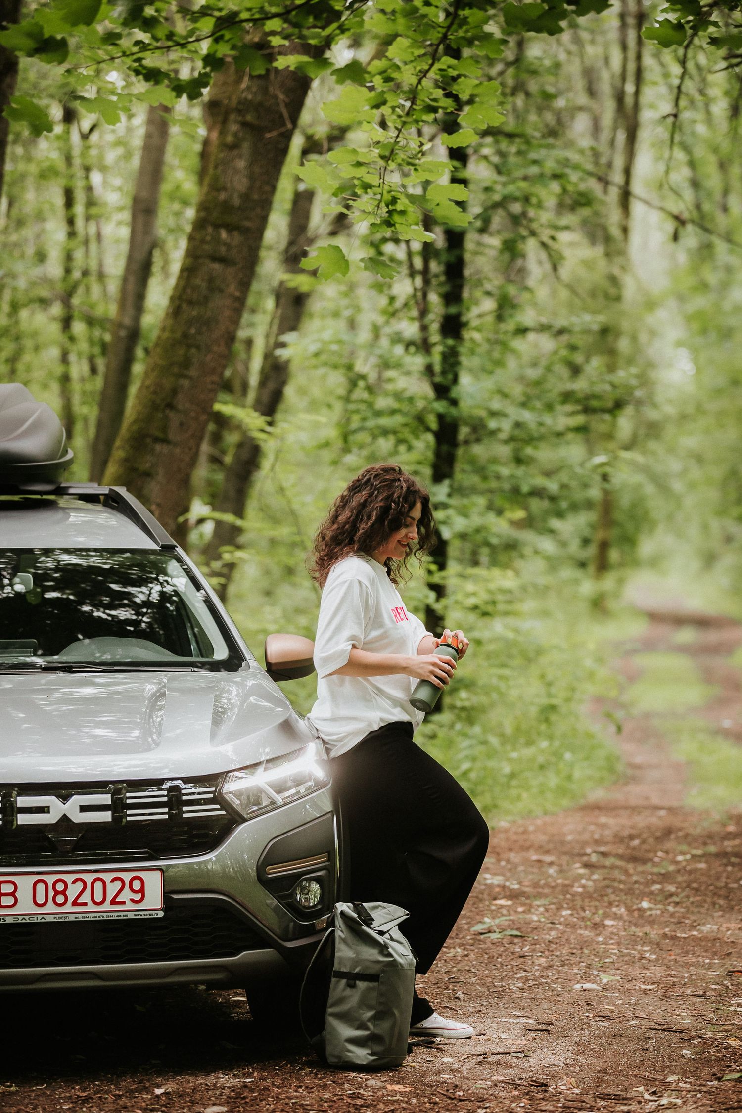 Woman leaning against a Dacia Duster SUV on a forest trail, wearing white top and black pants with a backpack nearby.