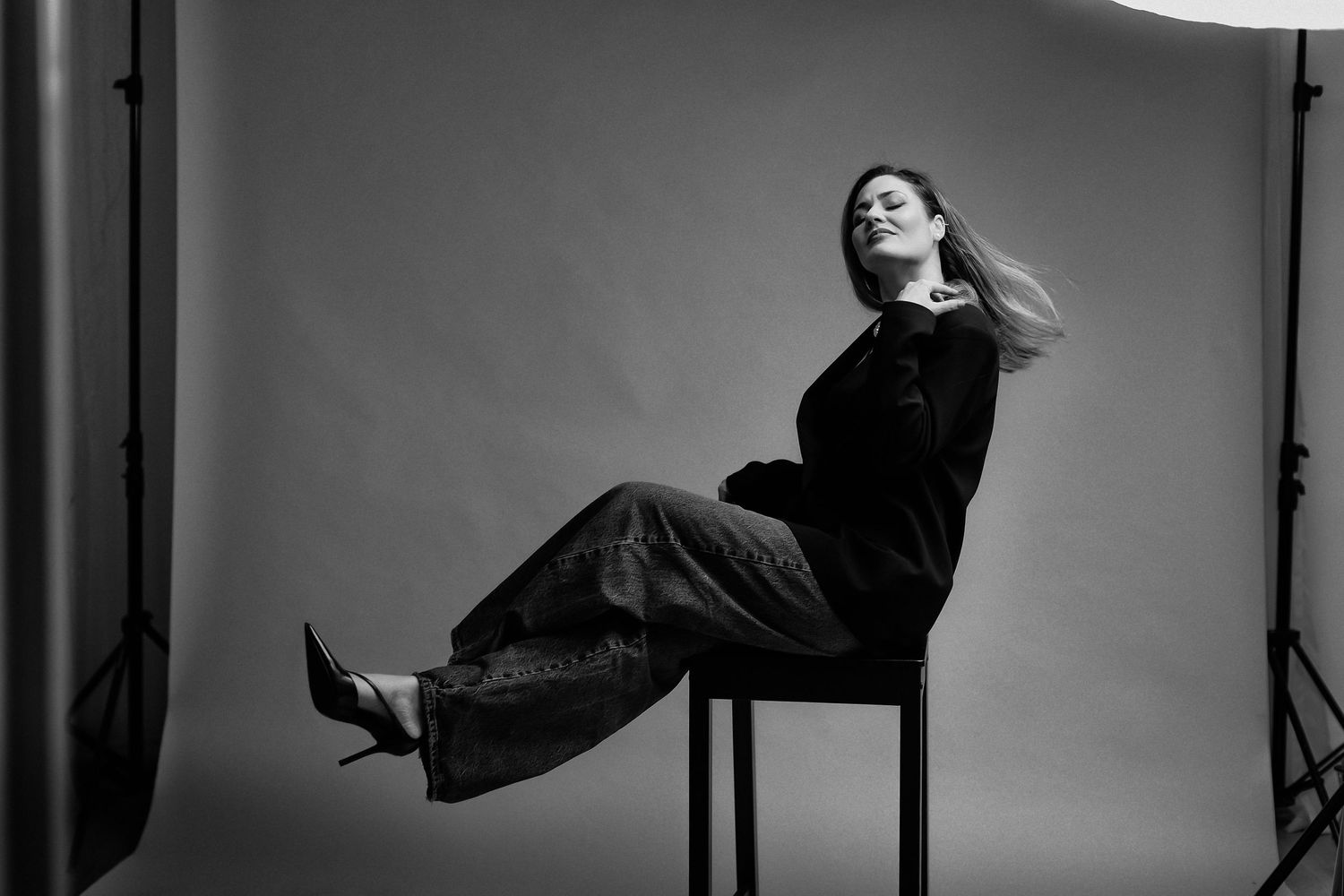 Black and white photo of a woman sitting on a stool, leaning back gracefully in dark wide-leg pants and blazer.