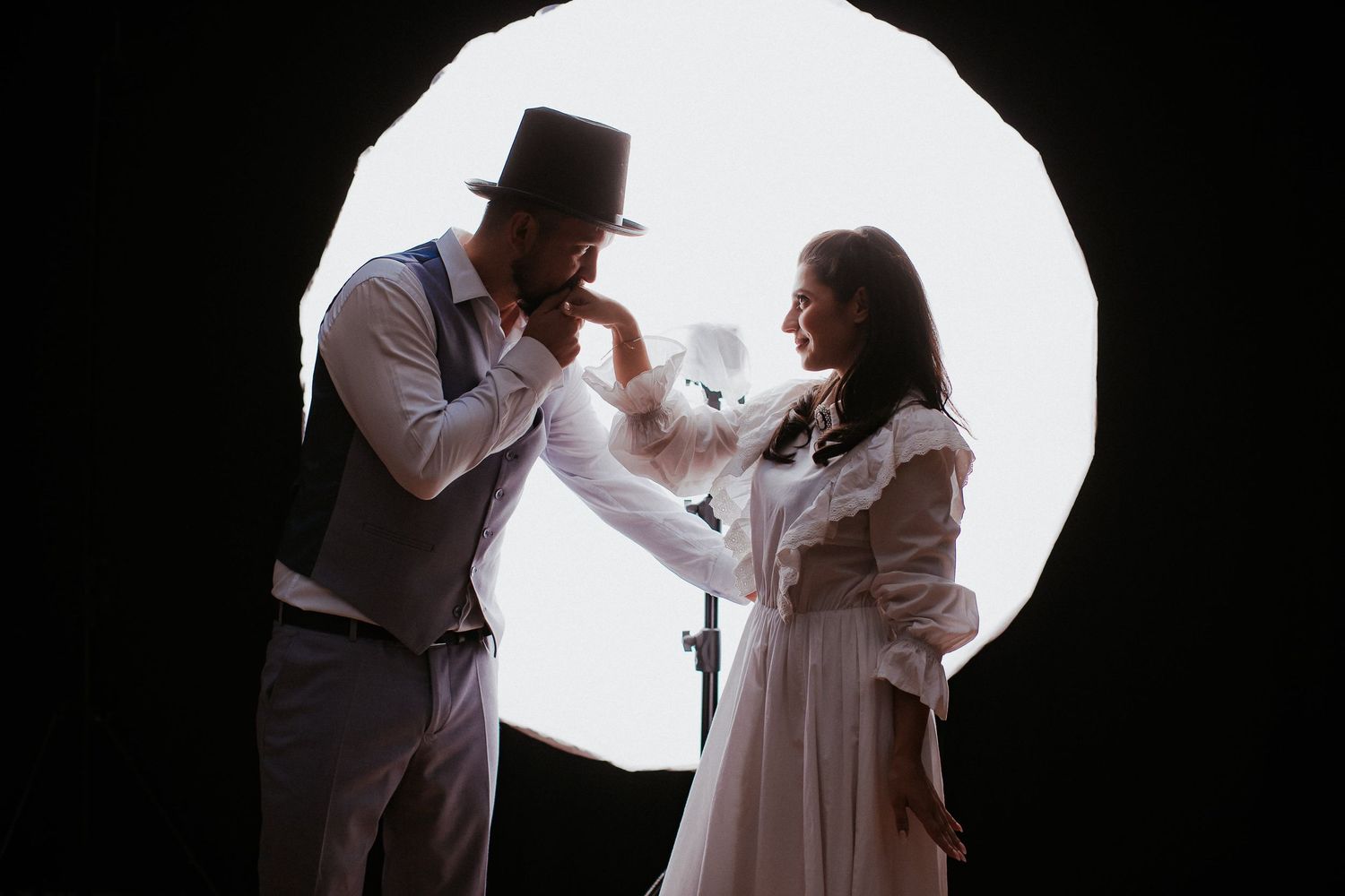 A man in a top hat kisses a woman's hand in a romantic vintage-styled portrait against a circular light.