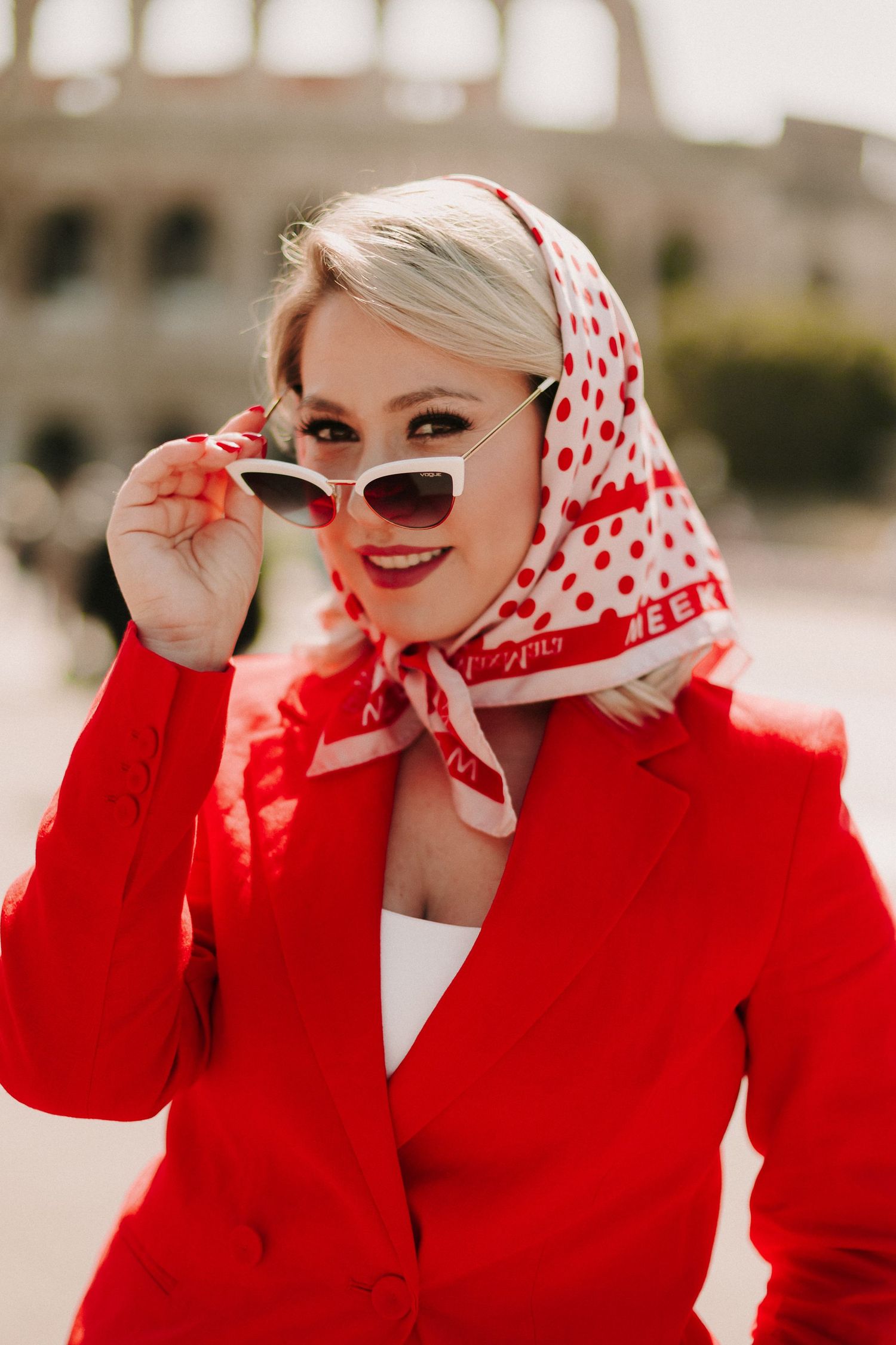 Stylish blonde woman in red blazer and polka dot headscarf lowering white sunglasses near the Colosseum.