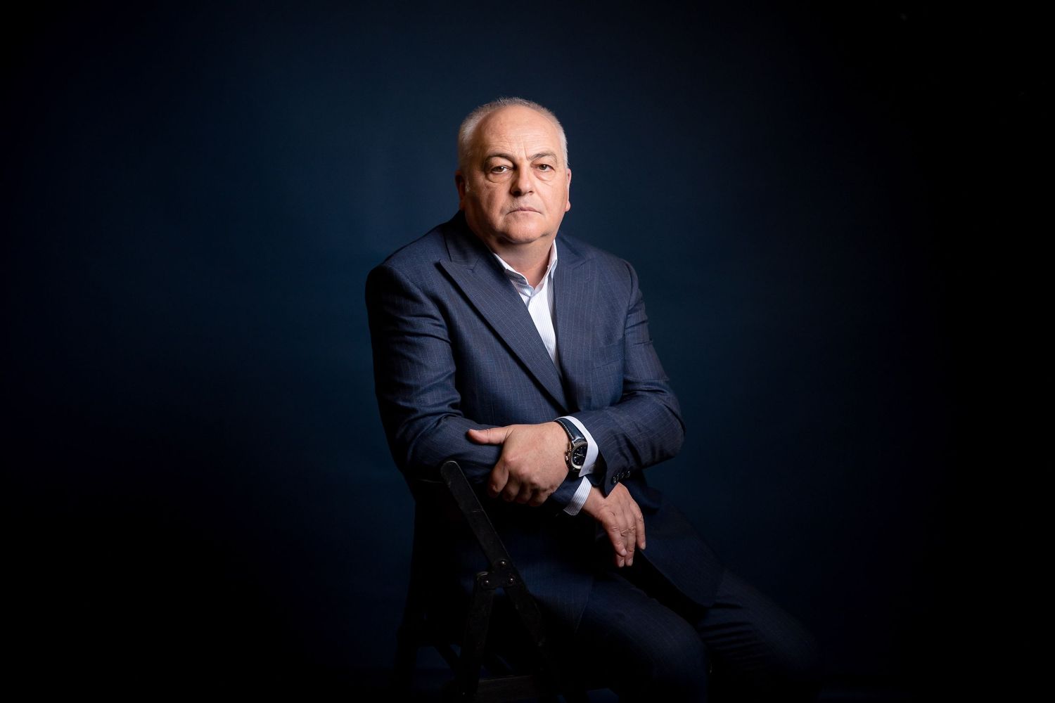 Professional businessman in navy suit seated confidently against dark blue background in dramatic studio portrait.