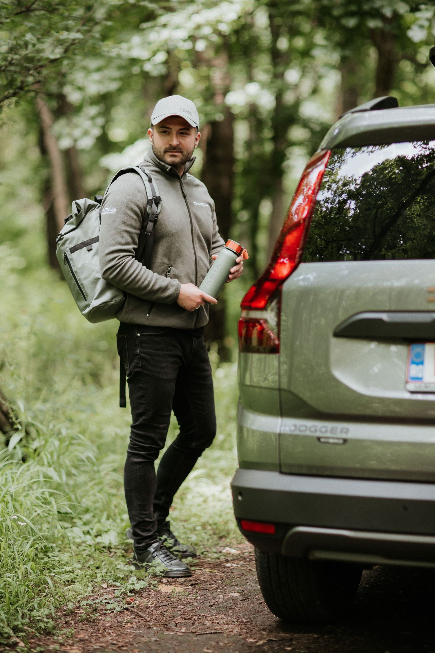 Man with backpack and thermos standing by a Dacia Jogger SUV in a lush green forest trail.