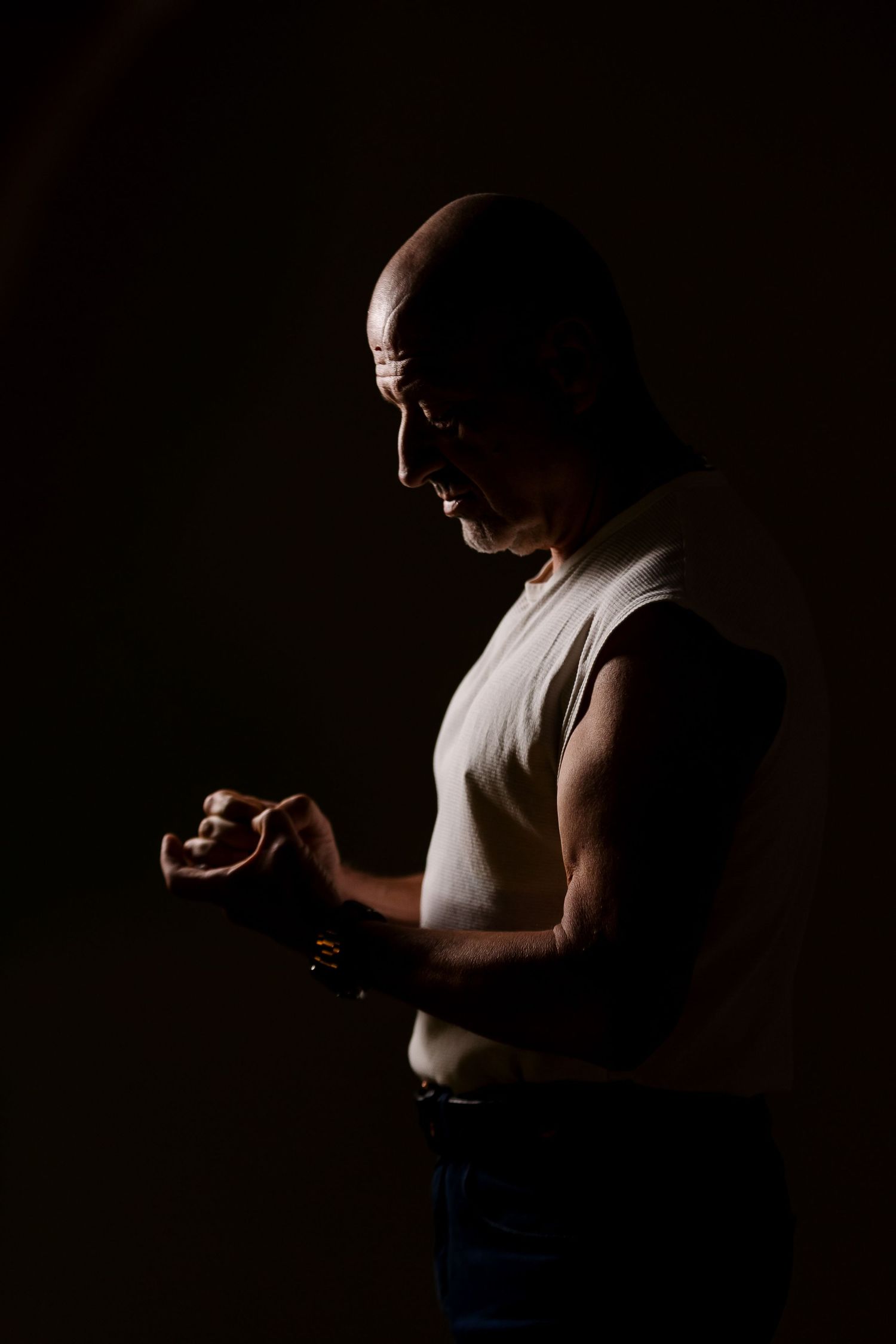 Bald muscular man in sleeveless shirt wrapping hands in dark dramatic studio lighting, side profile view.