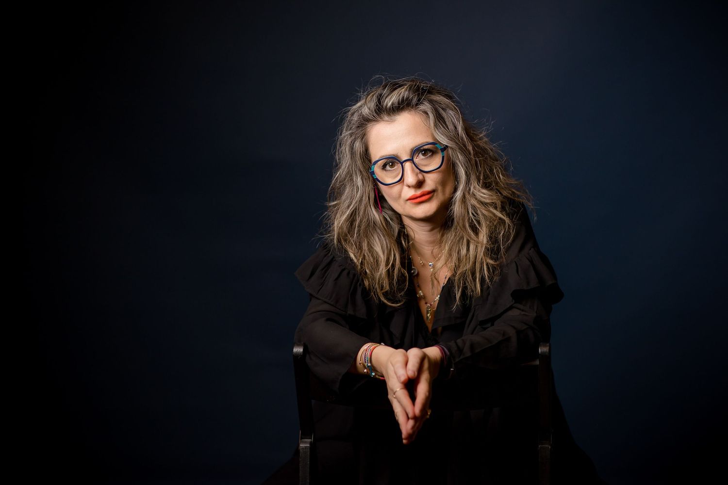 Woman with wavy hair and red lipstick wearing glasses and black jacket, posed thoughtfully against dark background.