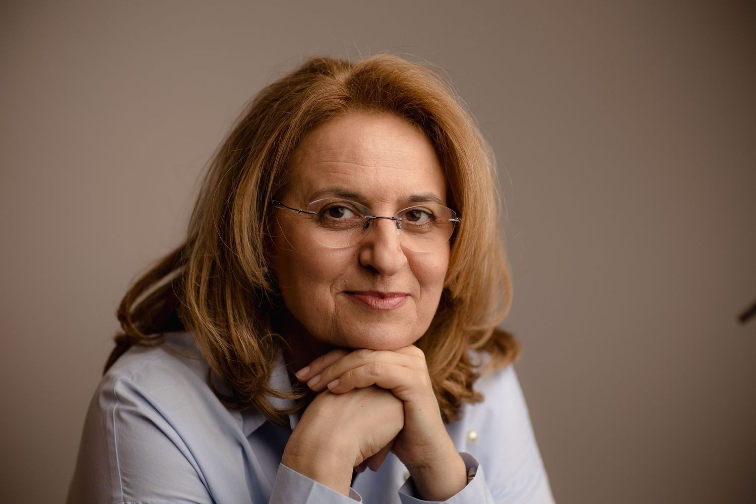 Professional woman with glasses and auburn hair smiling warmly, wearing a light blue shirt in a studio setting.