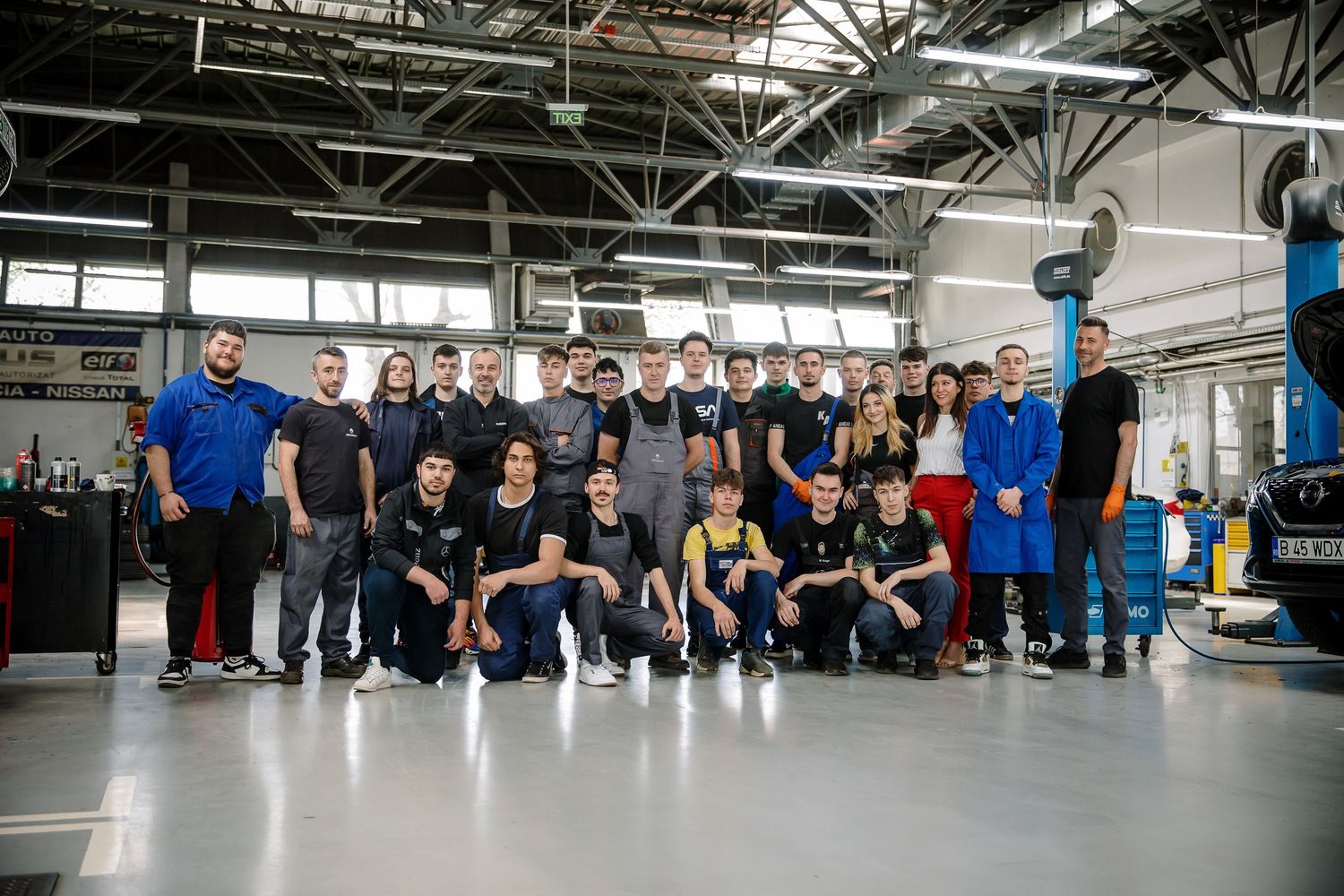 Large group of auto mechanics and students posing together inside a modern automotive repair workshop.