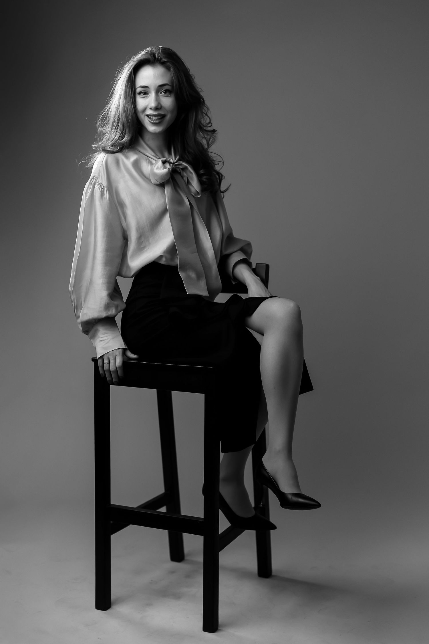 Black and white portrait of a smiling woman in blouse and skirt sitting elegantly on a bar stool.