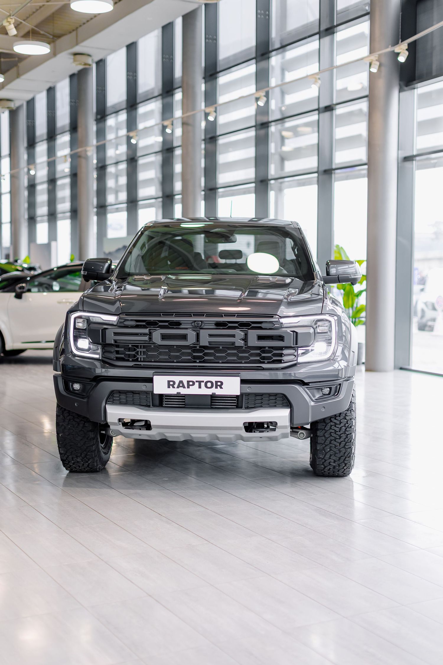 Dark gray Ford Raptor pickup truck displayed in a modern dealership showroom with large windows and white flooring.