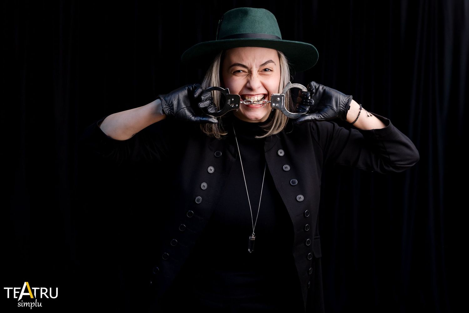 Woman in black outfit and green hat playfully biting gloves against dark background in theatrical pose.
