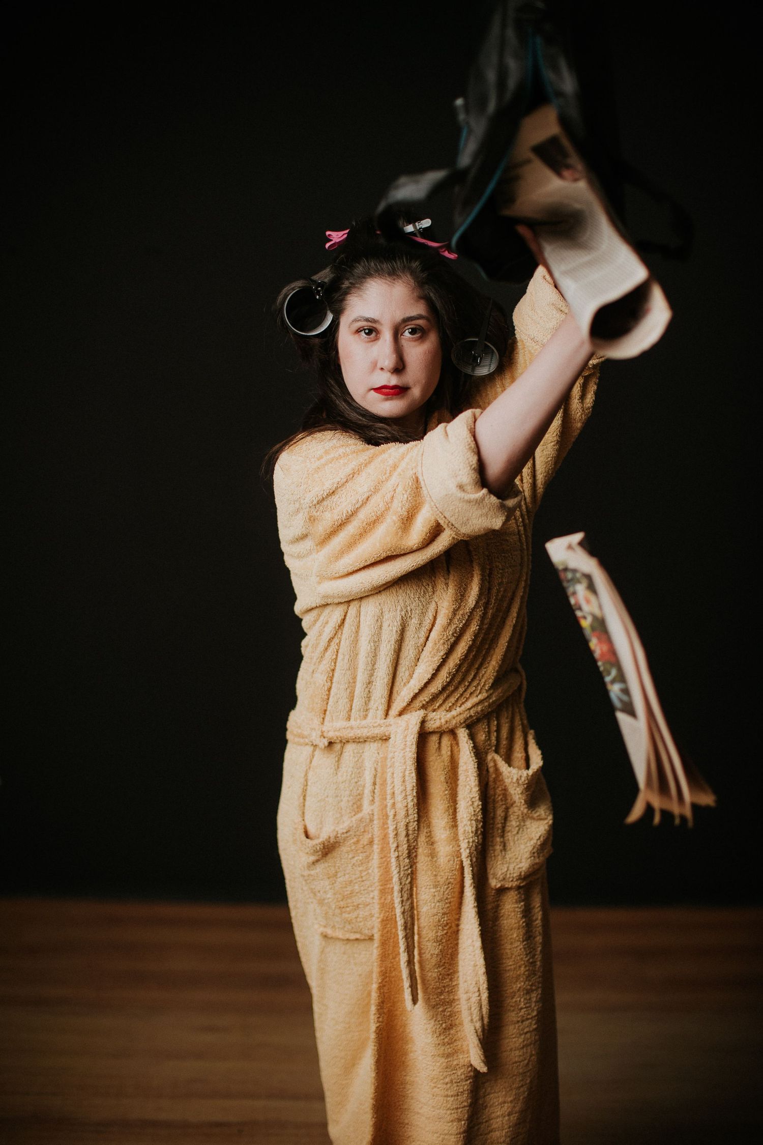 Woman in yellow robe and hair curlers holds up a handbag overhead against dark background.