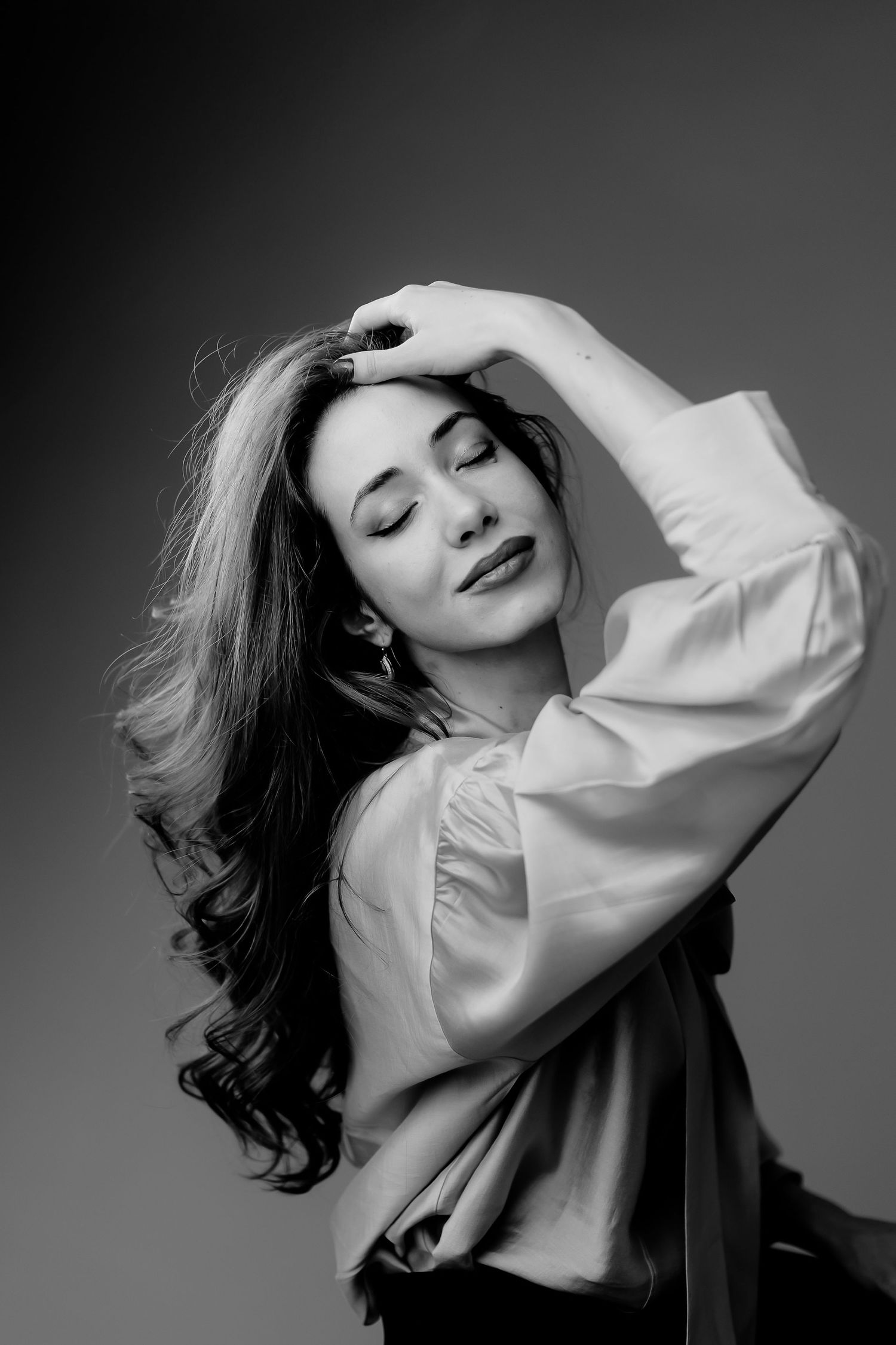 Black and white portrait of a woman with long wavy hair, eyes closed, hand raised gracefully above her head.