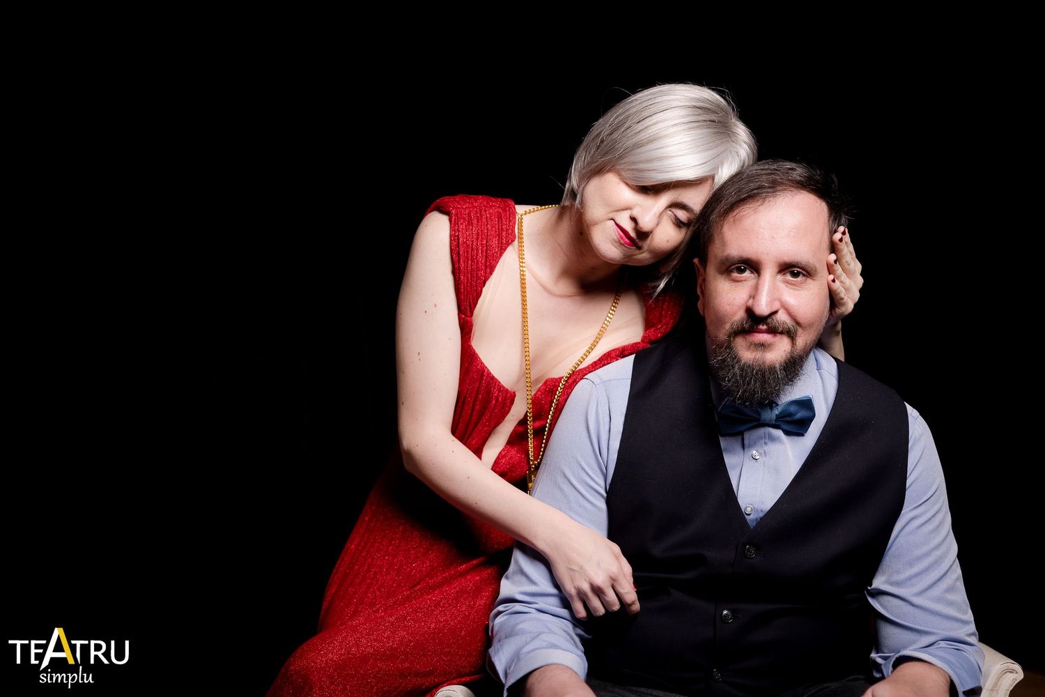 A woman in a red dress leans on a bearded man in a vest, posing for a professional theater portrait.