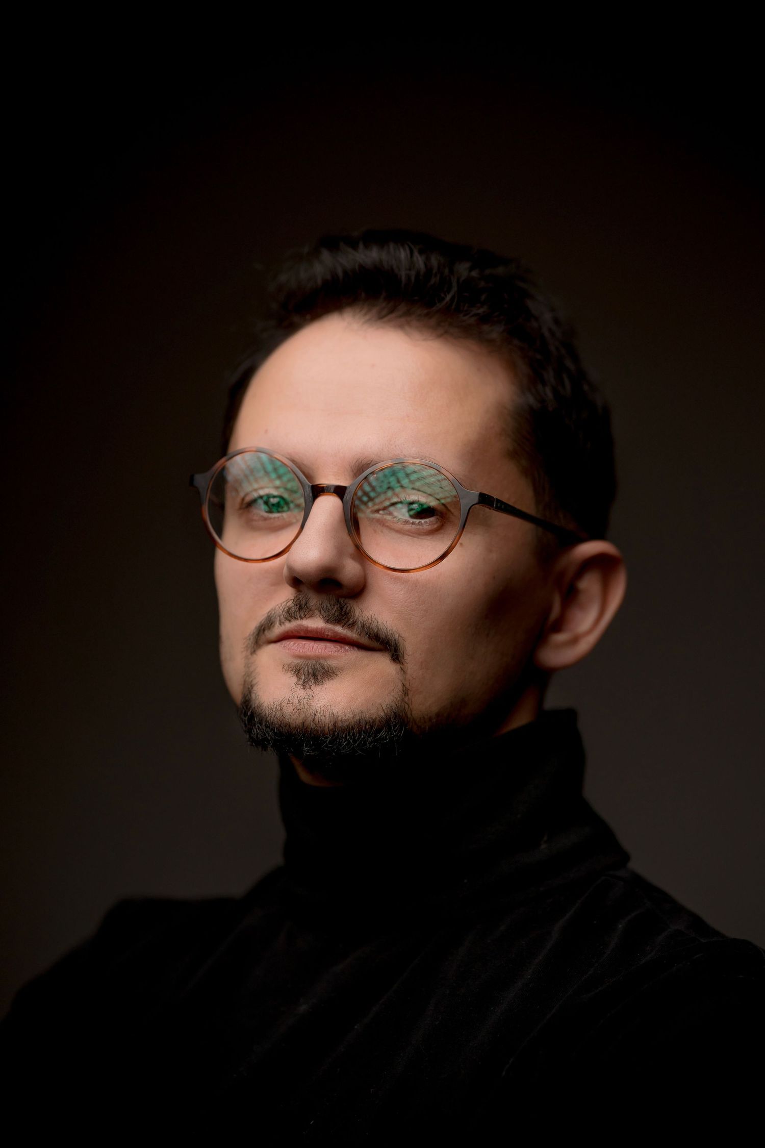 Professional portrait of a man with glasses, short dark hair, beard, wearing a black turtleneck sweater against dark background.