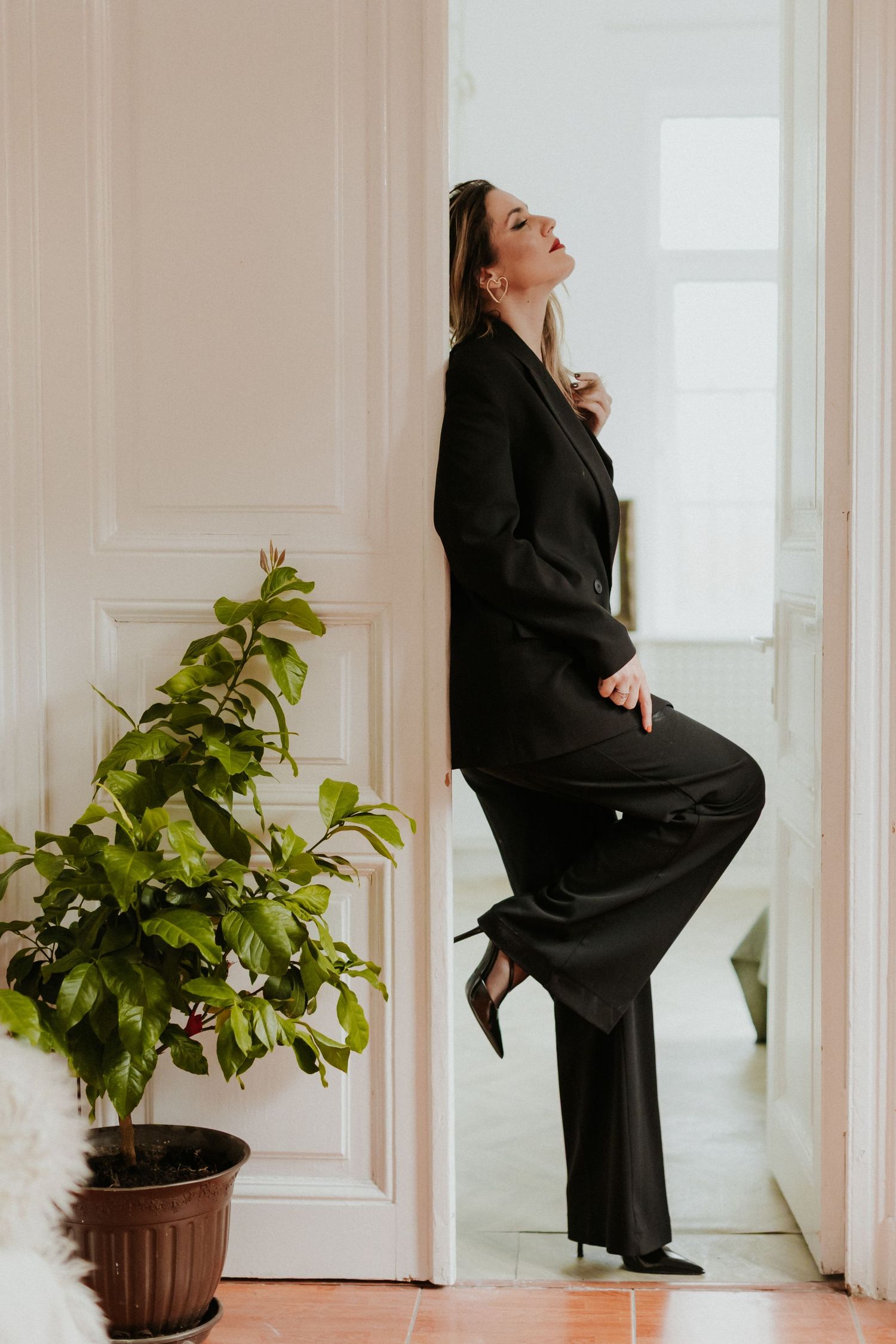 Woman in black suit posing against white door frame next to green potted plant in bright interior.
