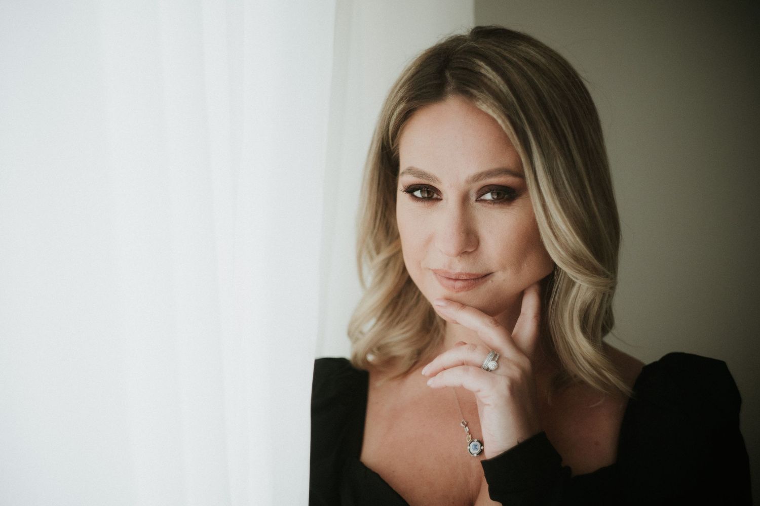 Elegant blonde woman in black top posing thoughtfully by window with soft natural light, wearing diamond ring.