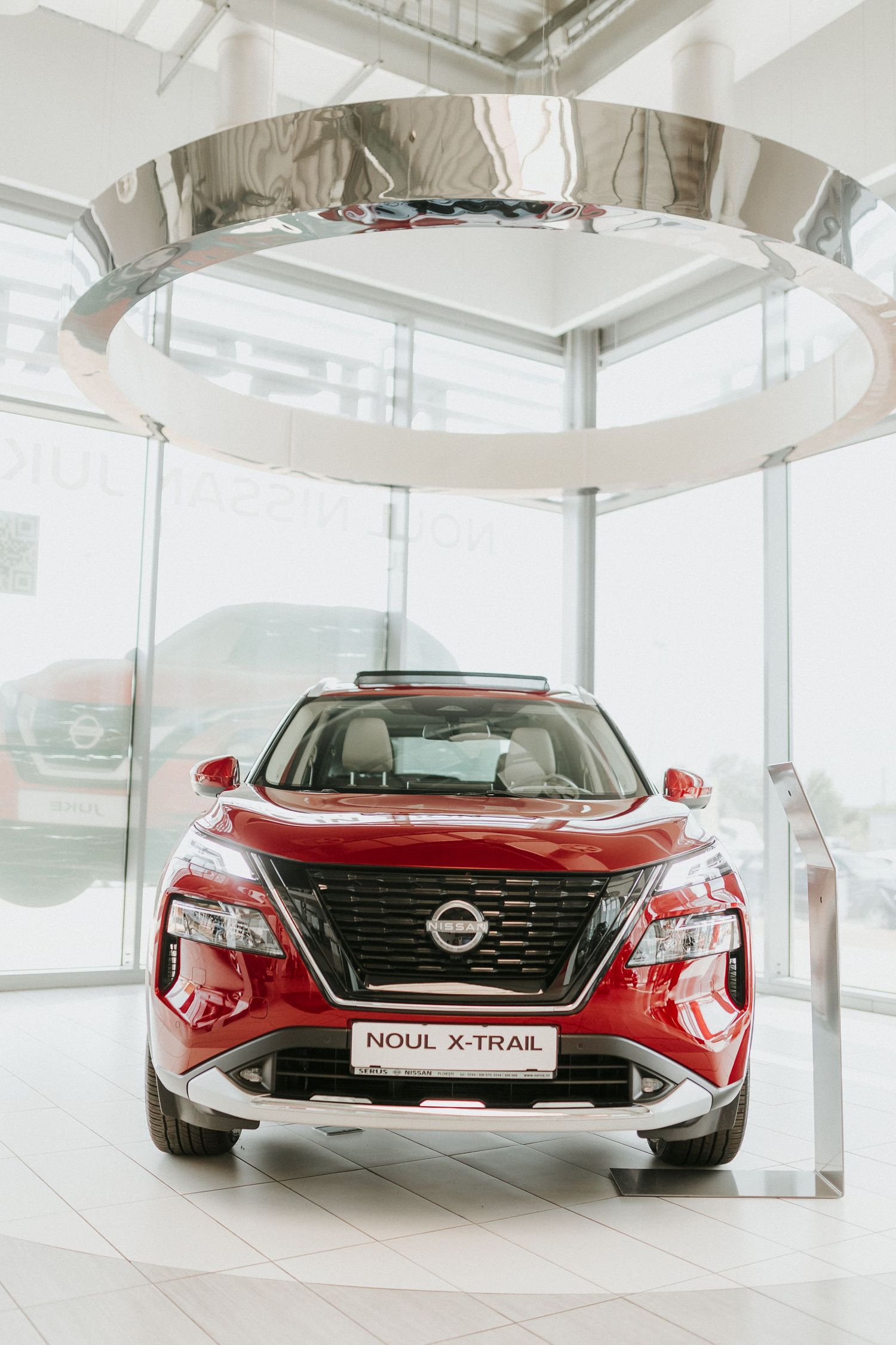 Red Nissan X-Trail SUV on display in a modern dealership showroom with large windows and chrome ring lighting.