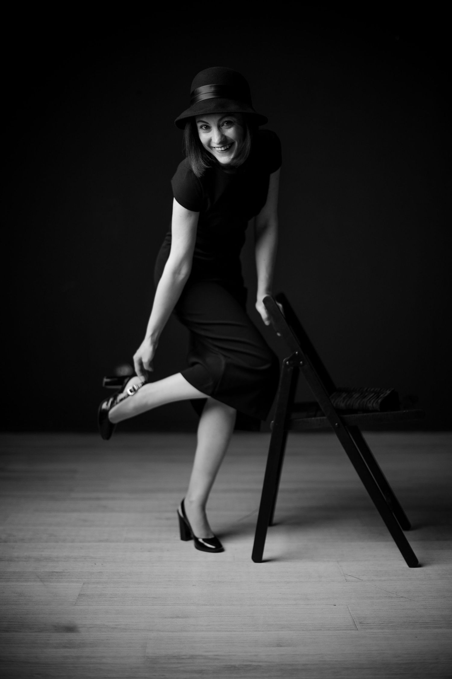 Black and white photo of a smiling woman in a black dress and hat, posing playfully beside a wooden chair.