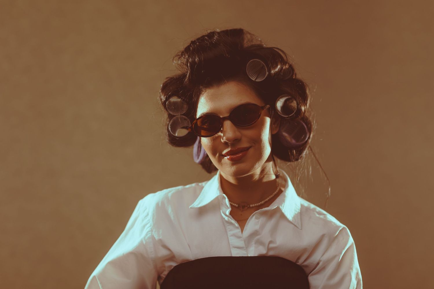 Woman with hair rollers and retro sunglasses smiling in a stylish white shirt against a warm background.