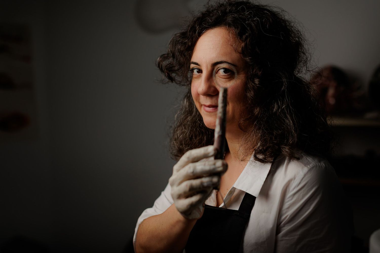 Woman in gloves and apron holds a tool, smiling in a dimly lit workshop setting.