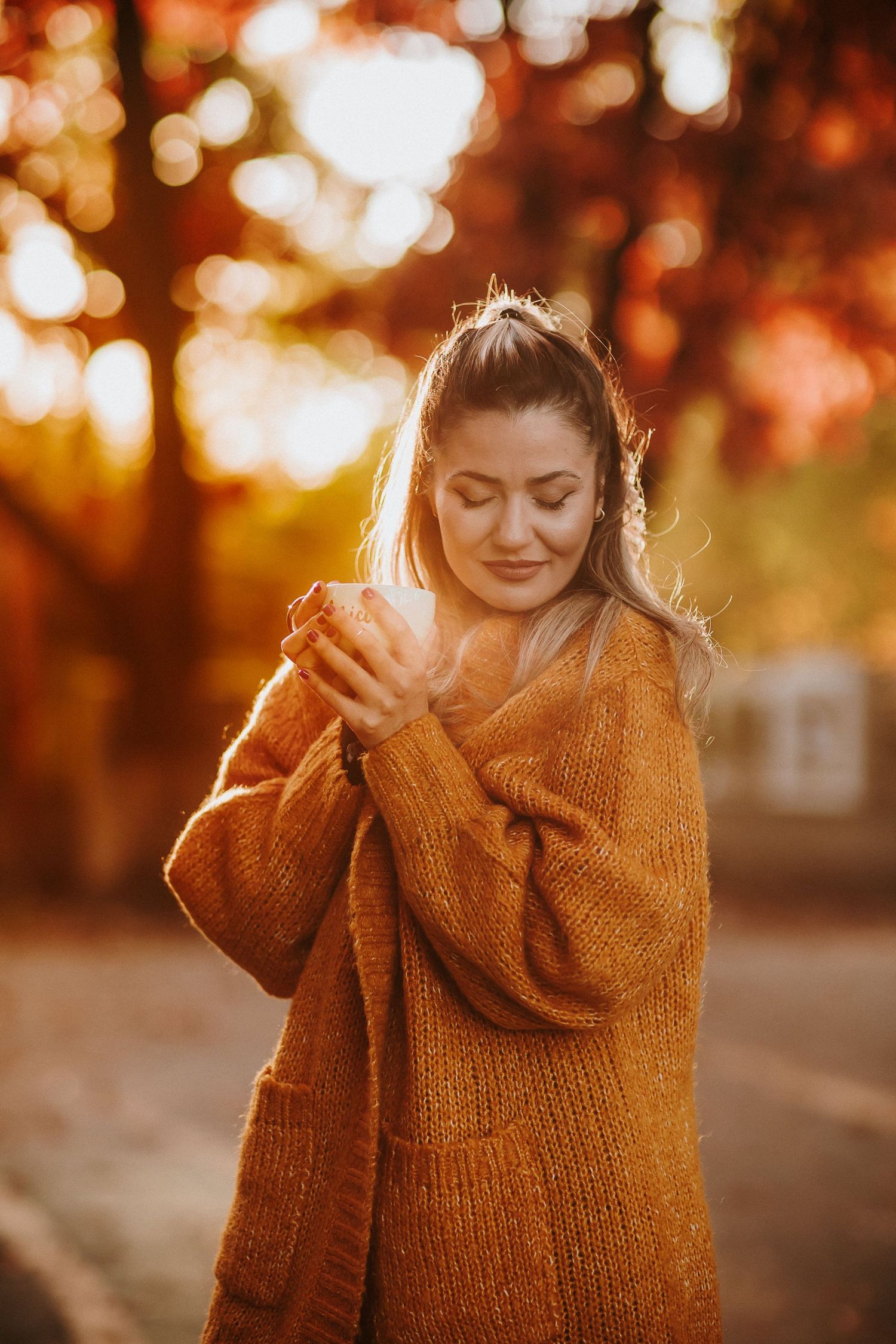 Woman in orange knit sweater holding a warm drink outdoors, surrounded by vibrant autumn foliage in golden sunlight.