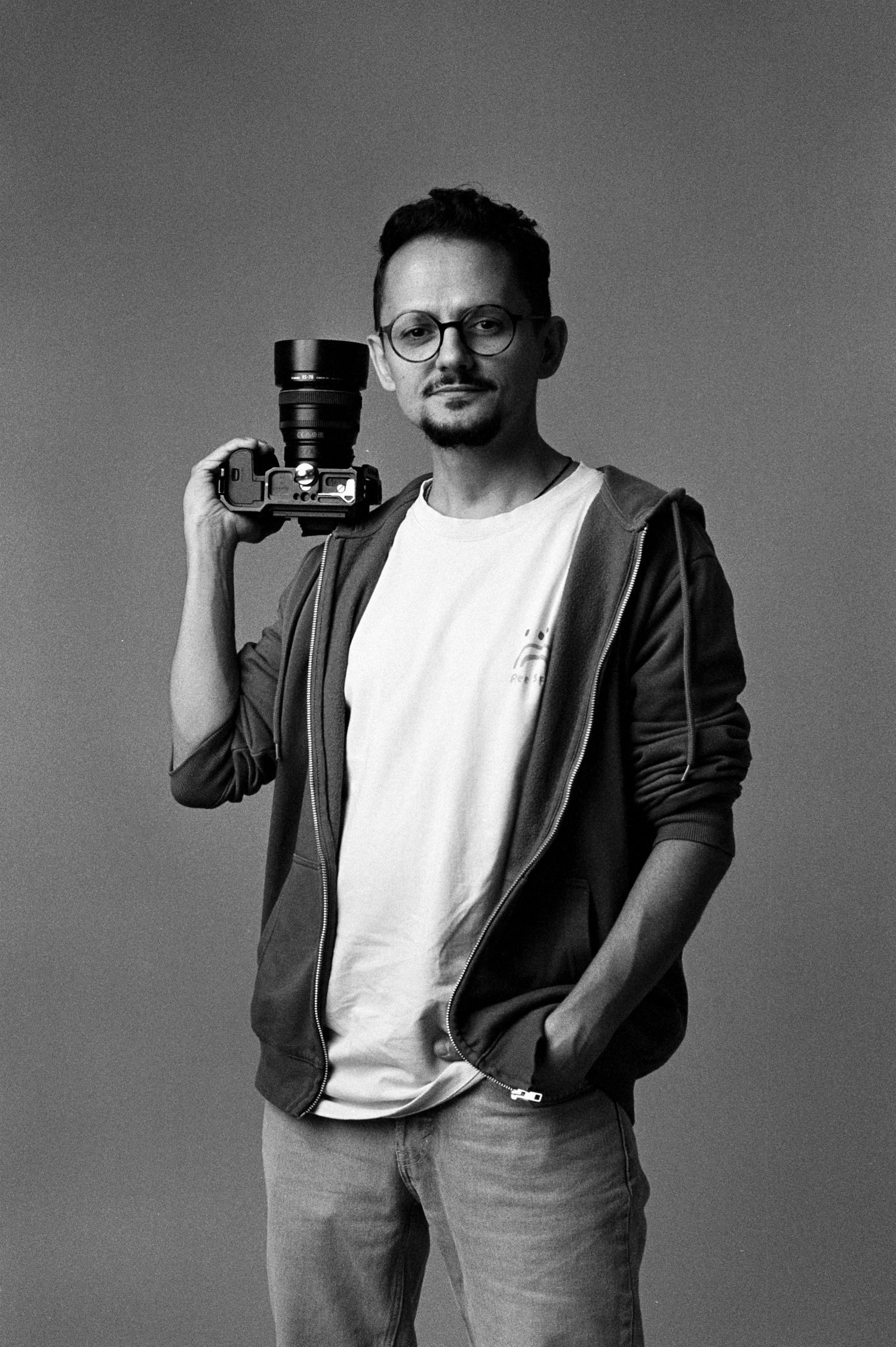 Black and white portrait of a young man with glasses holding a camera on his shoulder against a gray background.