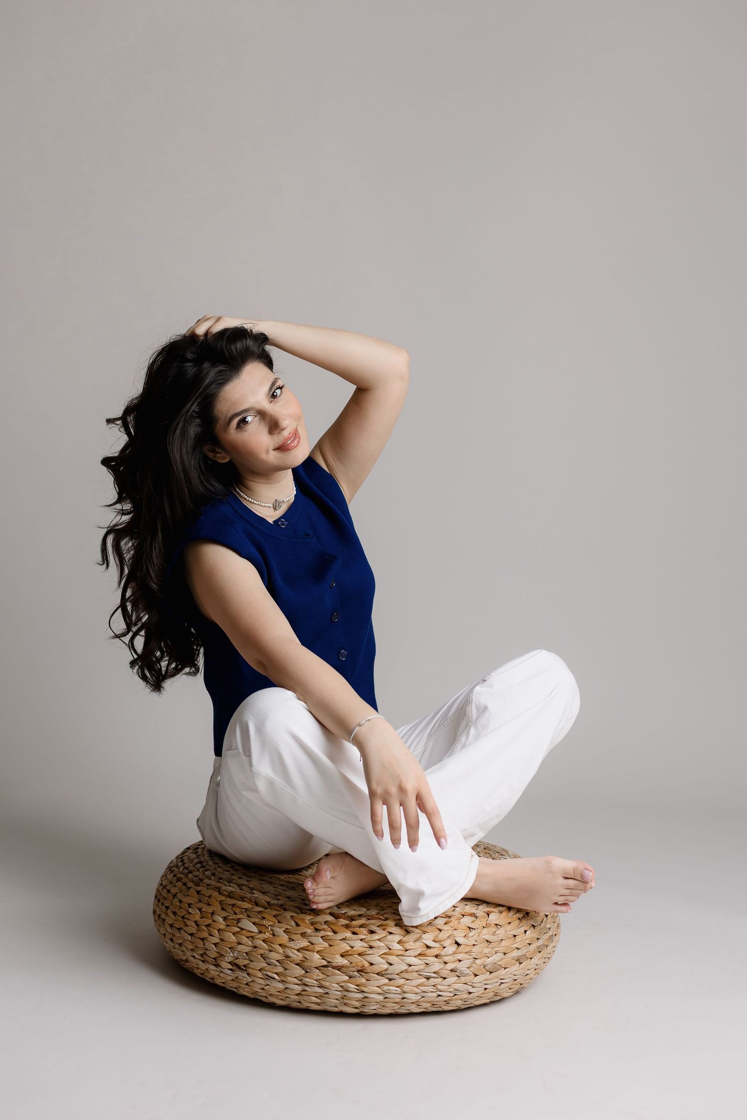 Woman in blue top and white pants sitting cross-legged on woven pouf, posing with hand in dark hair.