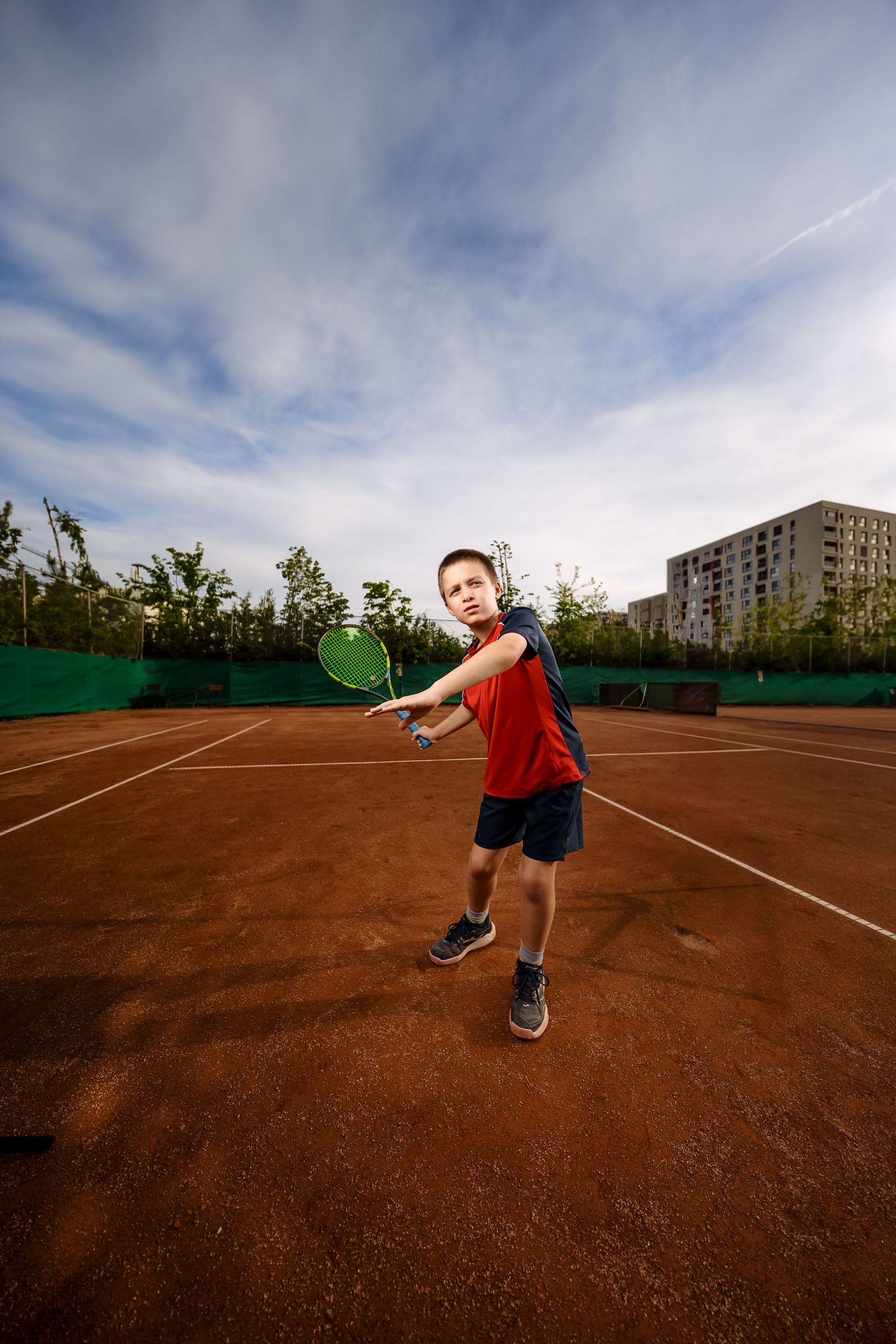 Young boy in red shirt playing tennis on clay court, mid-swing with green racket under blue sky.