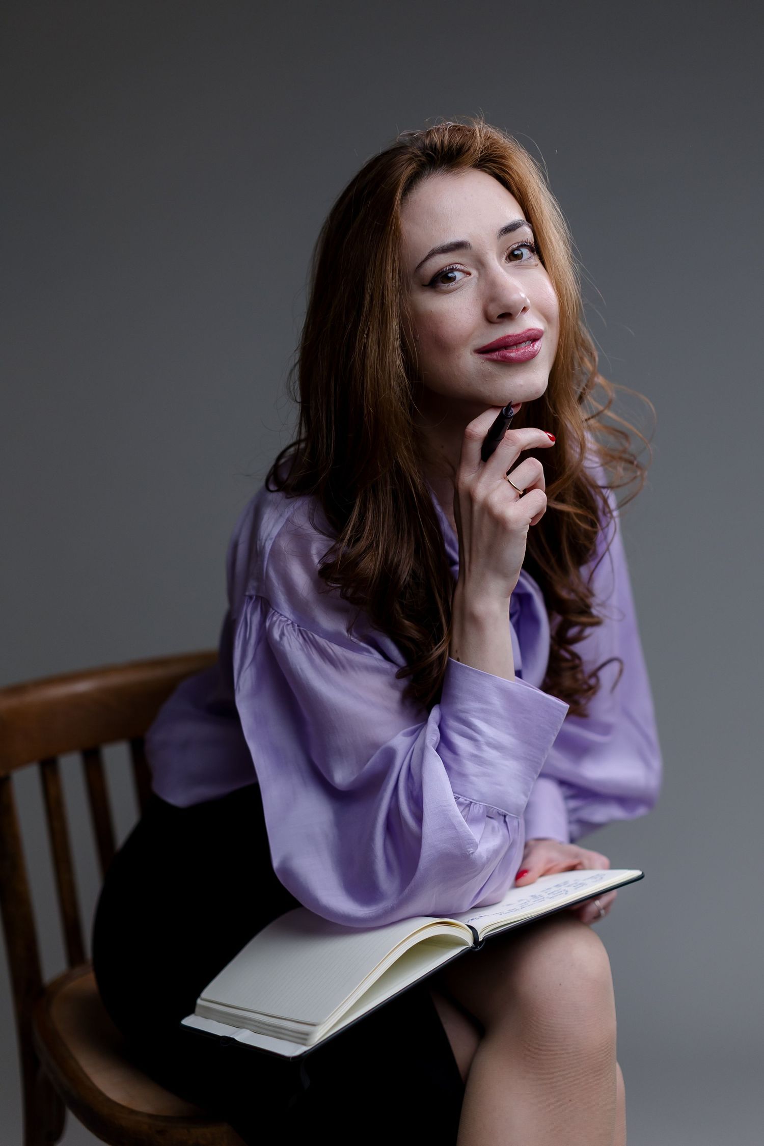 Young woman in purple blouse sits on wooden chair holding open notebook, smiling thoughtfully against gray background.