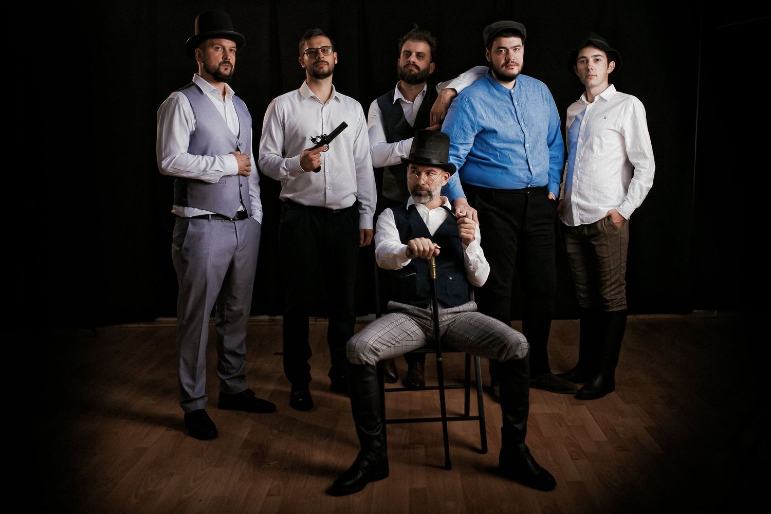 Six men in vintage Western attire pose dramatically in a dark studio, one seated holding a gun.