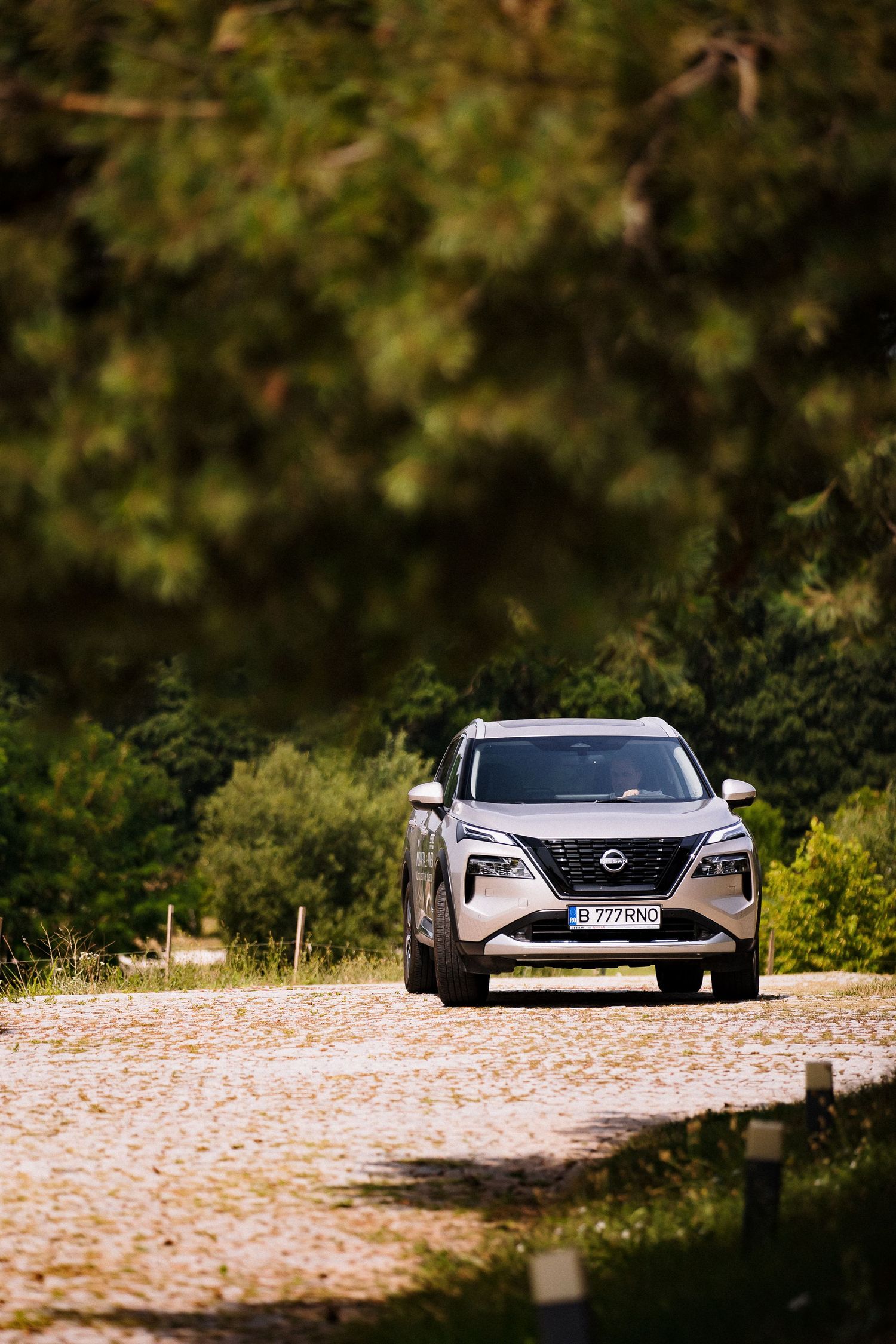 Silver Nissan X-Trail SUV driving on a cobblestone path surrounded by lush green trees.